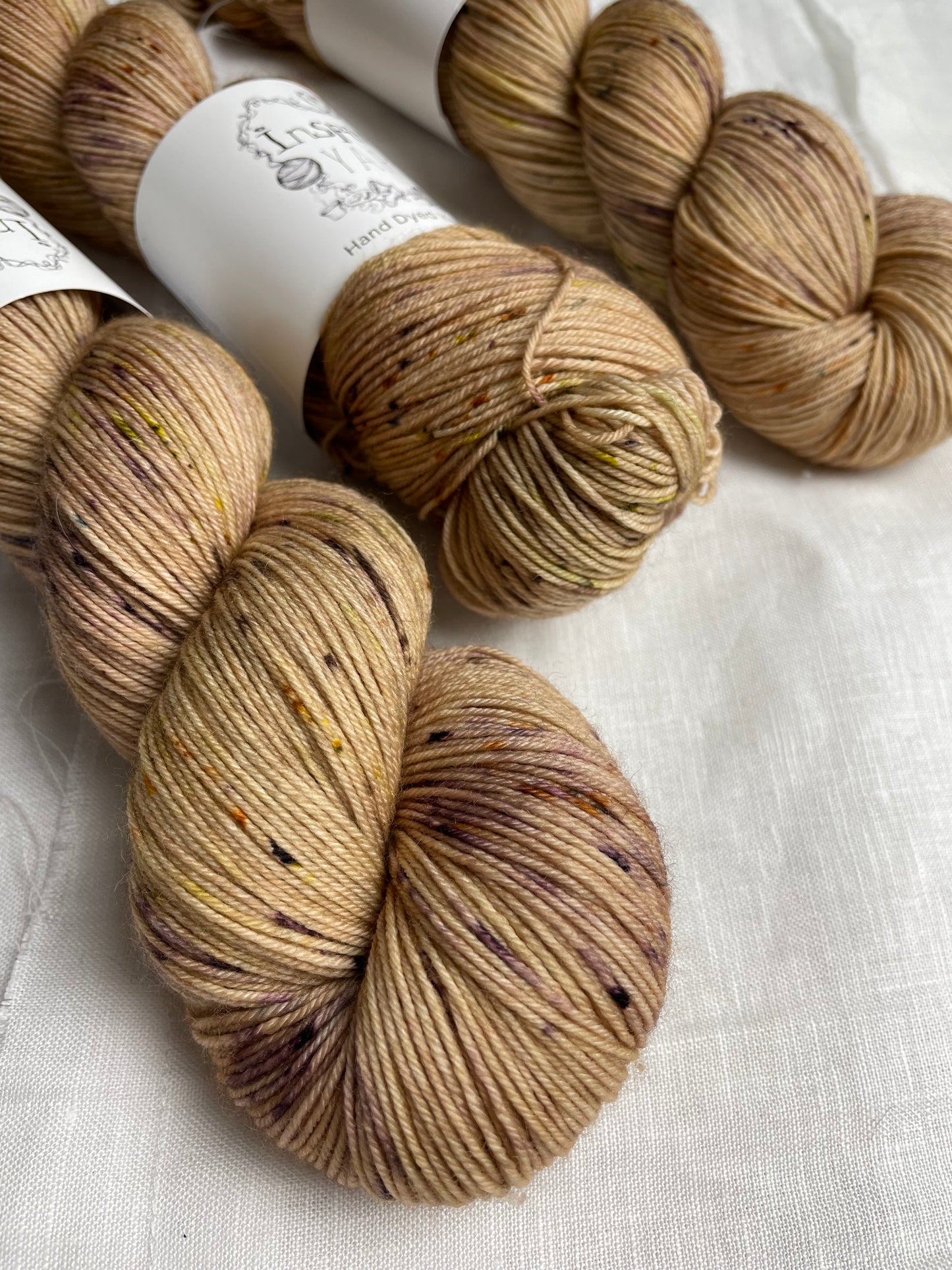 Close up of 3 skeins of Harvest, a pale gold with speckled coloured Merino Smooth Sock, labelled Inspiring Yarns on a white linen background.