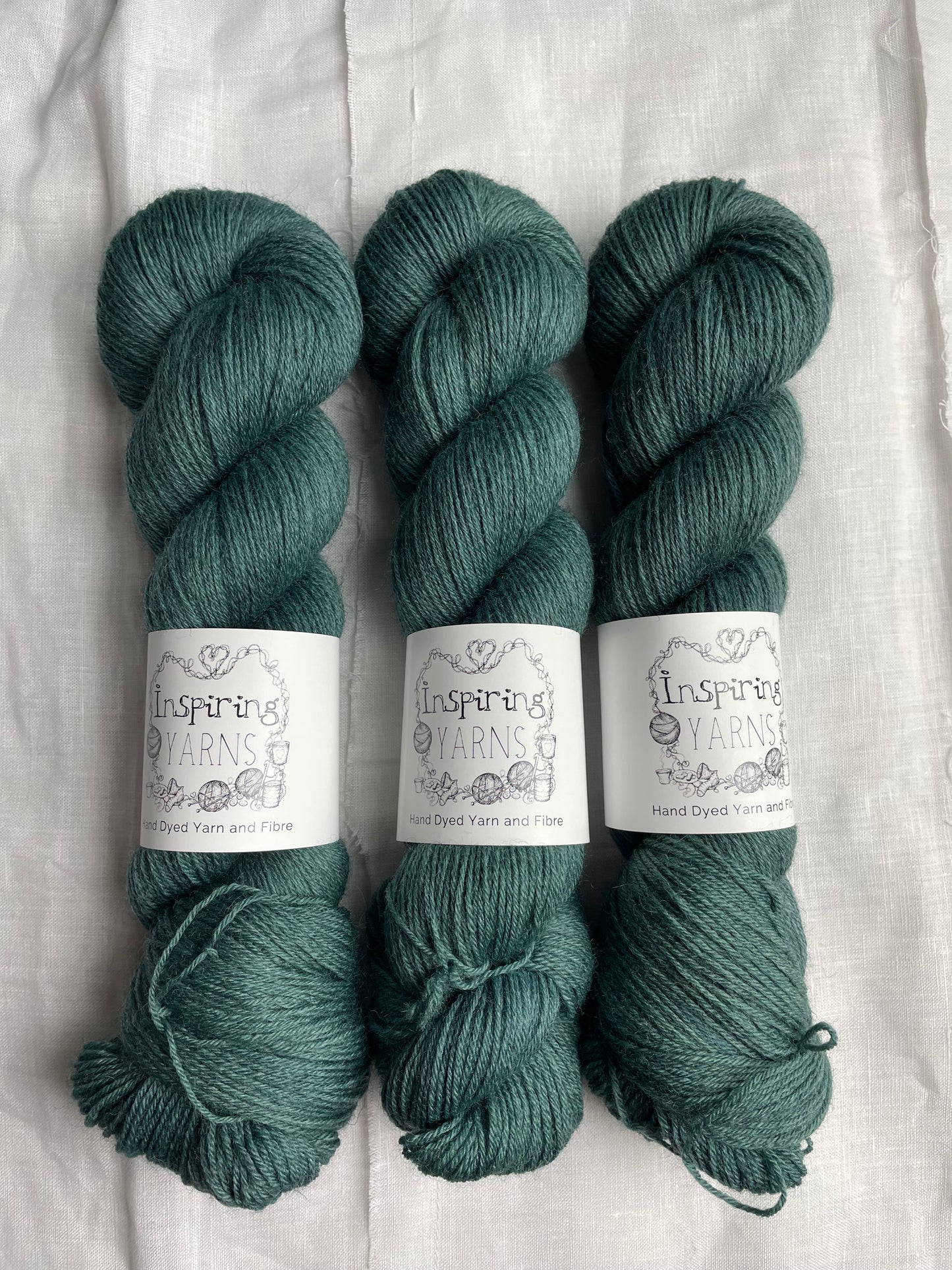 Three skeins of a tonal dark teal Highland 4Ply yarn branded Inspiring Yarns on a white linen background.