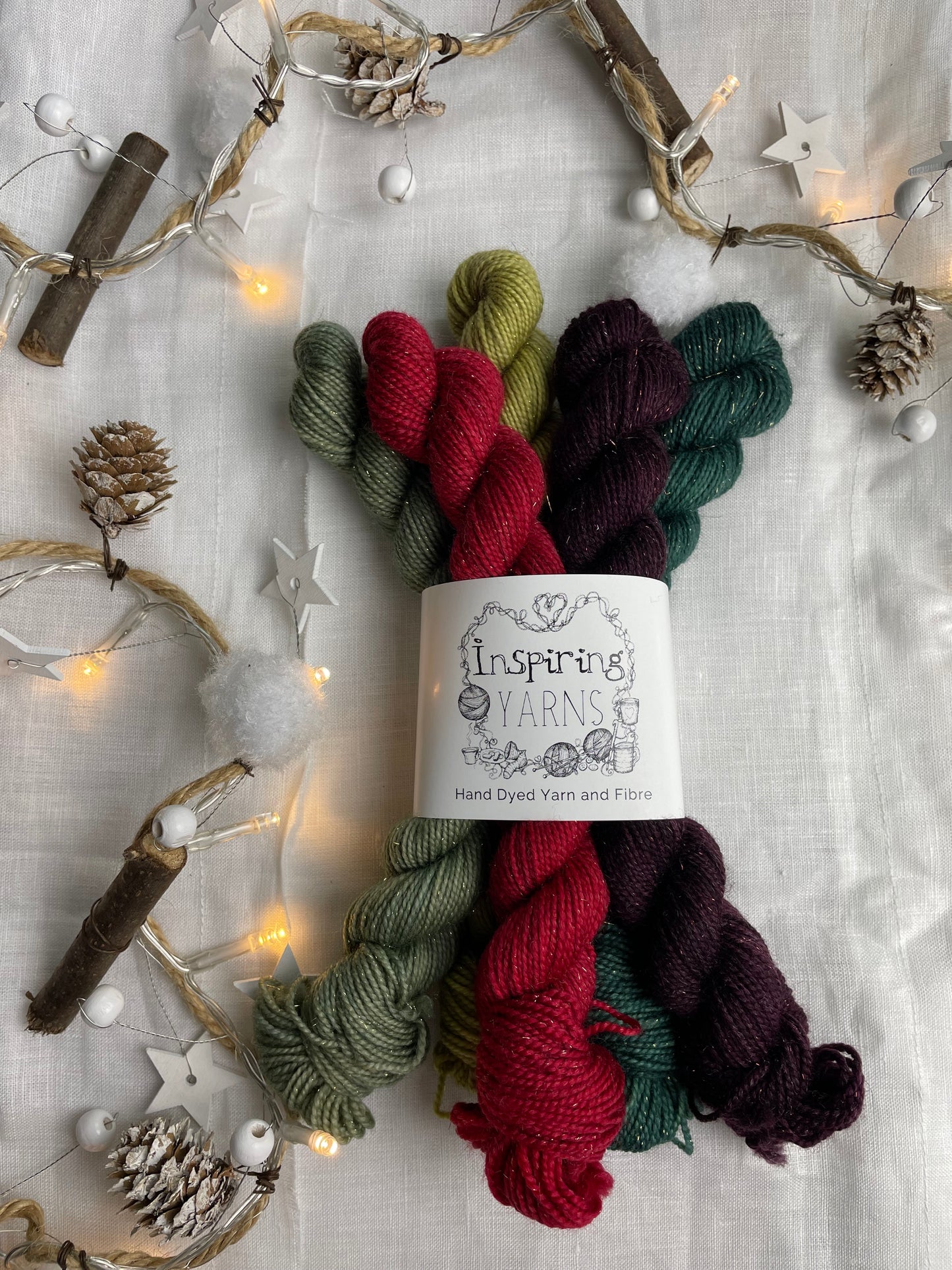 A set of five hand dyed yarn mini skeins on Sparkle Sock Yarn in red and green tones labelled Inspiring Yarns. The background is white linen and some Christmas lights with pinecones, twigs and stars.