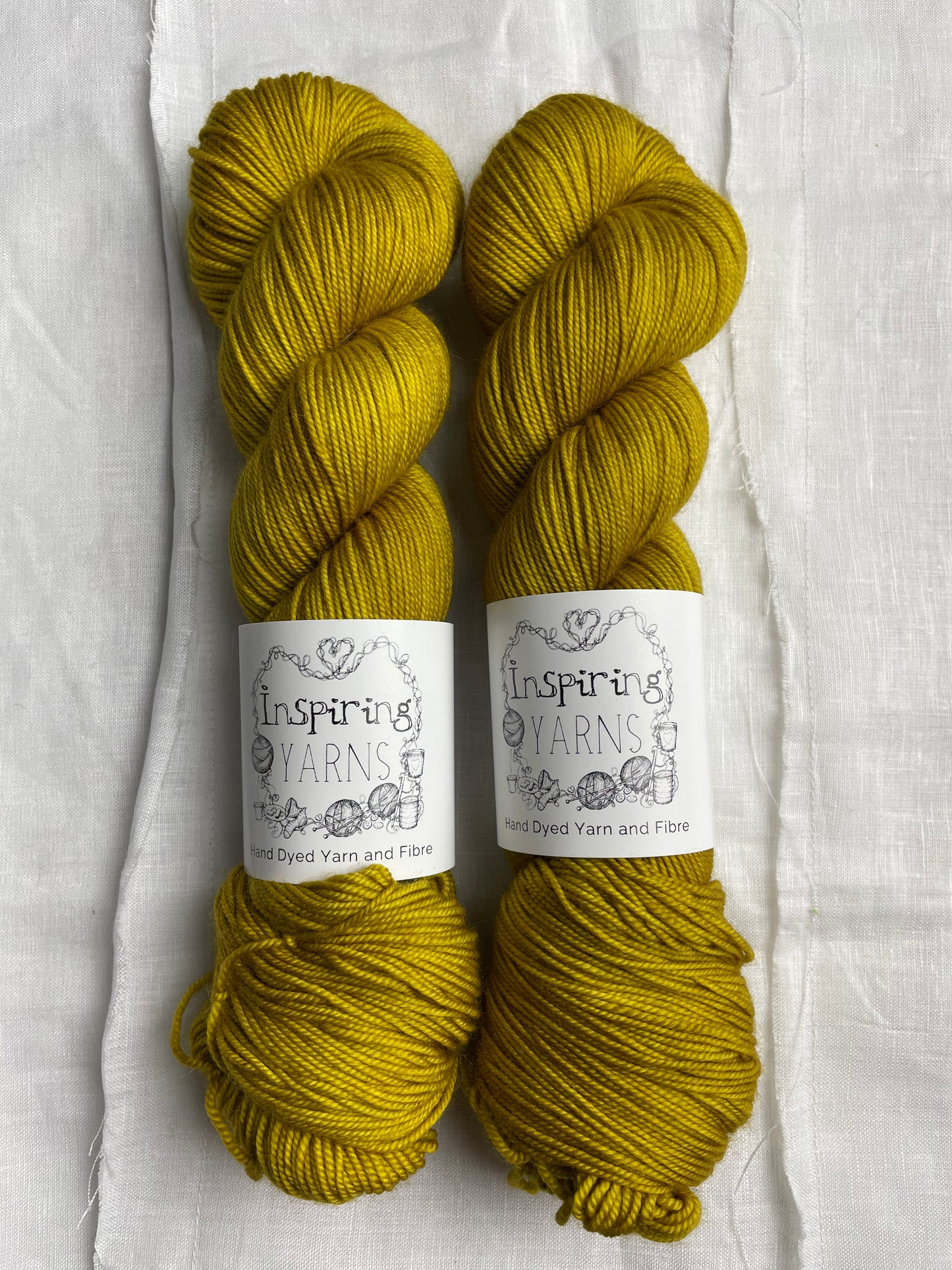 Two skeins of tonal golden ochre coloured hand dyed Merino Sport yarn branded Inspiring Yarns on a background of white linen.