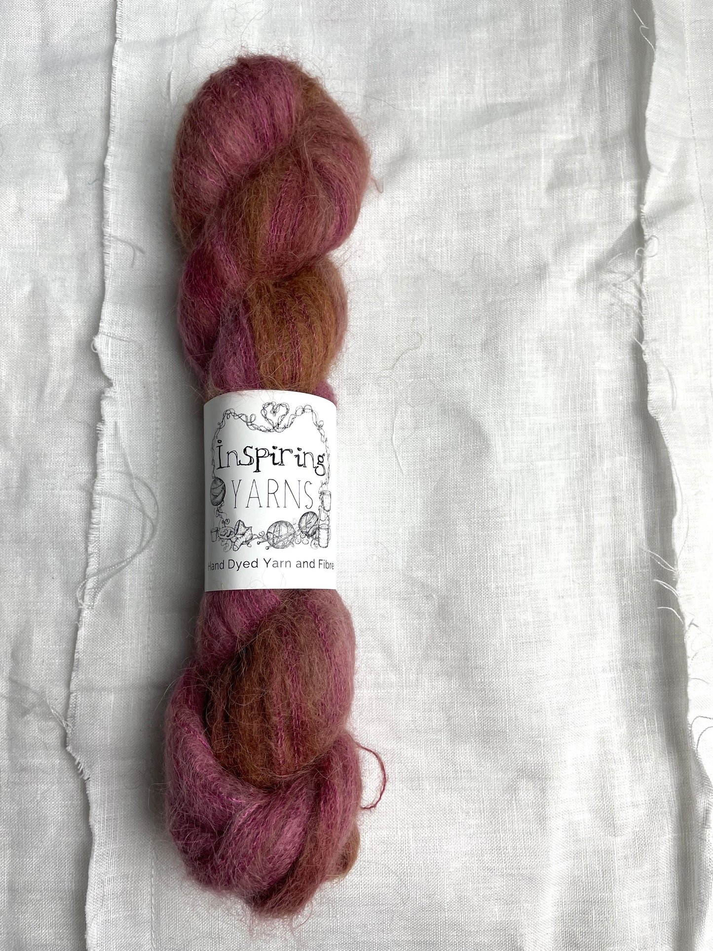 A skein of dusky pink Brushed Baby Suri Silk lace yarn labelled Inspiring Yarns on a white linen background.