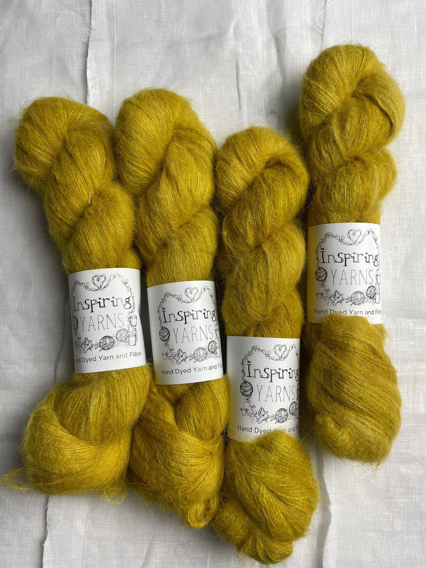 Four skeins of tonal golden ochre coloured hand dyed fluffy Alpaca Silk Lace yarn labelled Inspiring Yarns on a white linen background.