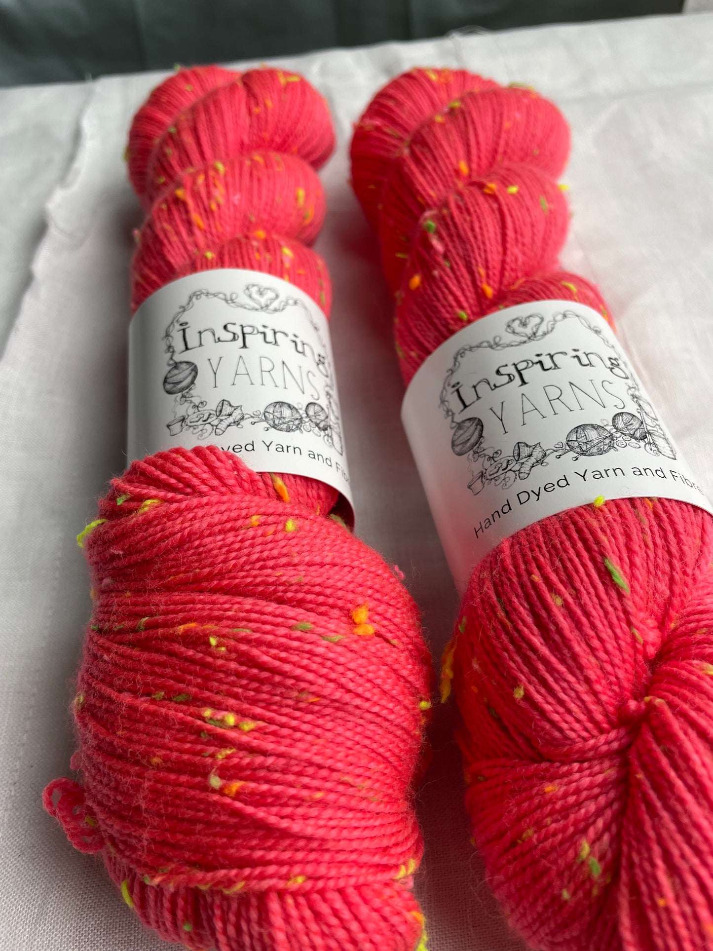 Close up of two skeins of tonal dark neon coral coloured hand dyed Disco Donegal yarn branded Inspiring Yarns on a white linen background. The yarn has multi coloured neon neps.