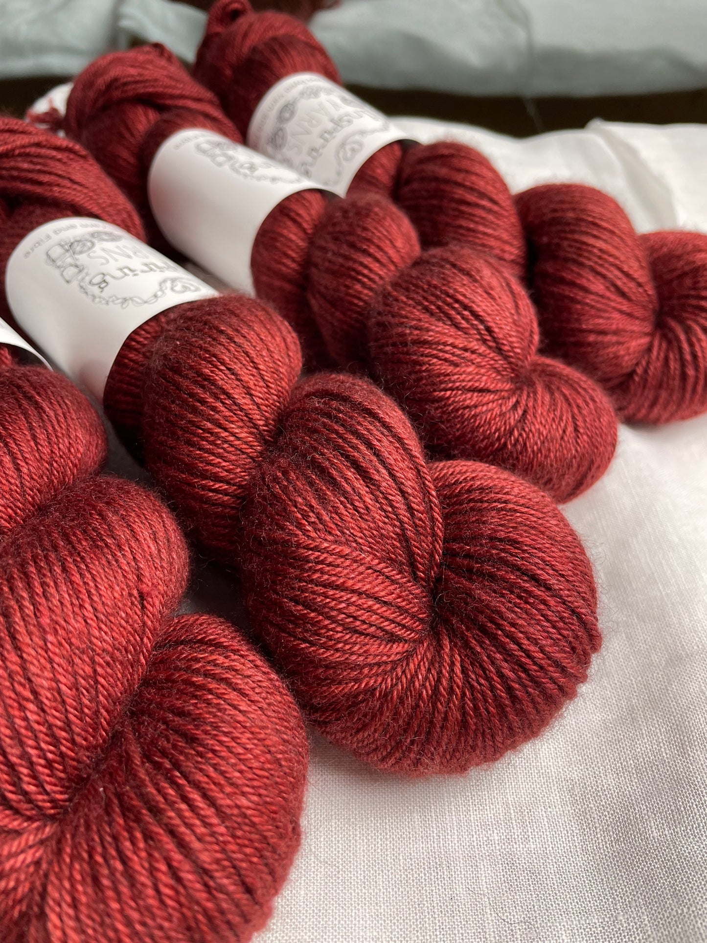 Close up of Four skeins of rusty red coloured yak silk dk yarn branded Inspiring Yarns on a white linen background.