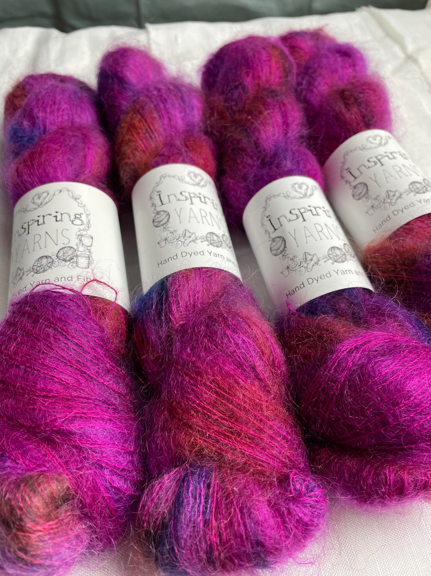 Close up of Four skeins of fuchsia pink speckled kid mohair silk lace yarn labelled Inspiring Yarns on a white linen background.