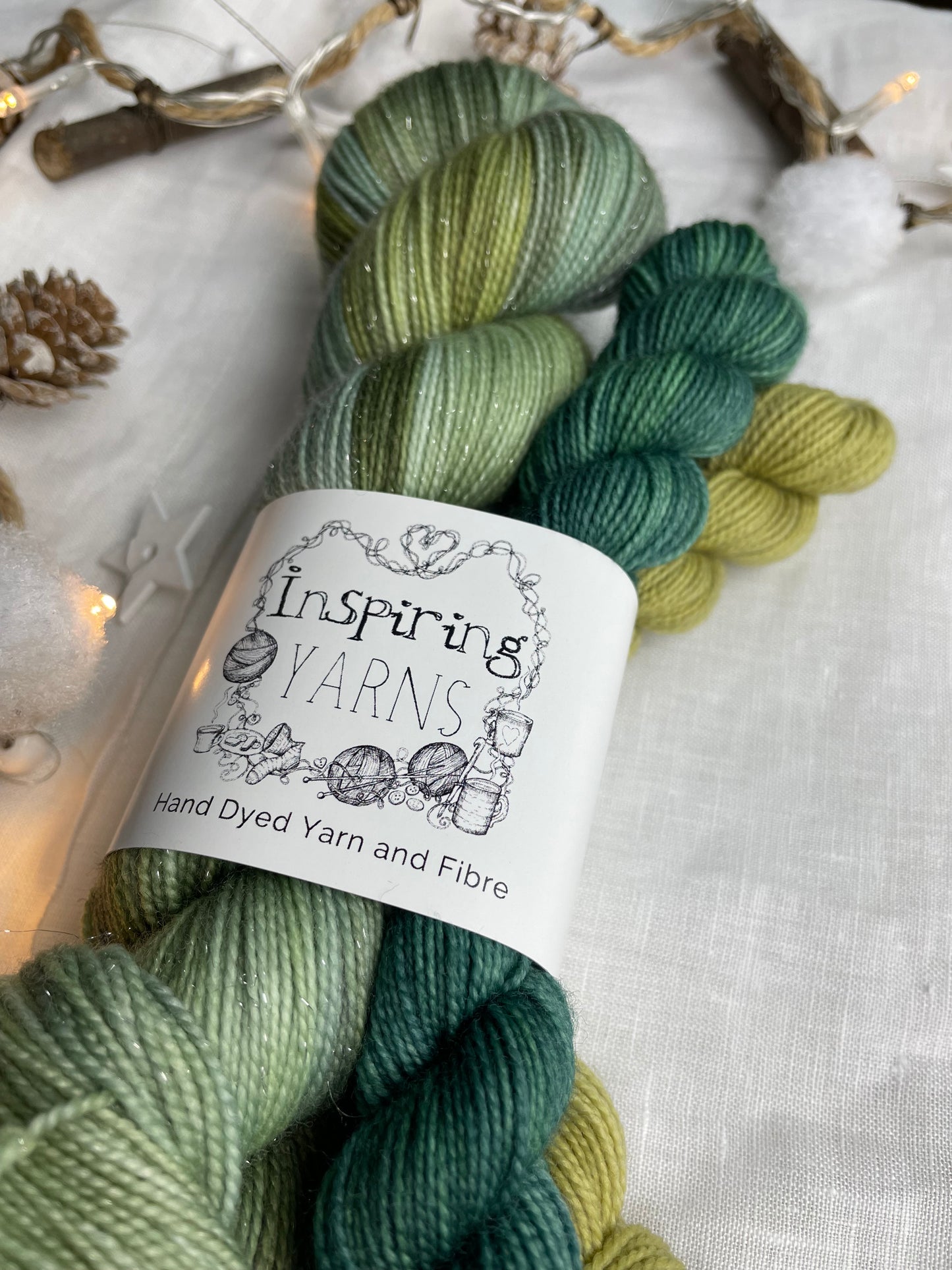 A hand dyed yarn sock set featuring large skein of Sparkle Sock Yarn in green tones with a teal and a lime green mini skein labelled Inspiring Yarns. The background is white linen and some Christmas lights with pinecones, twigs and stars.