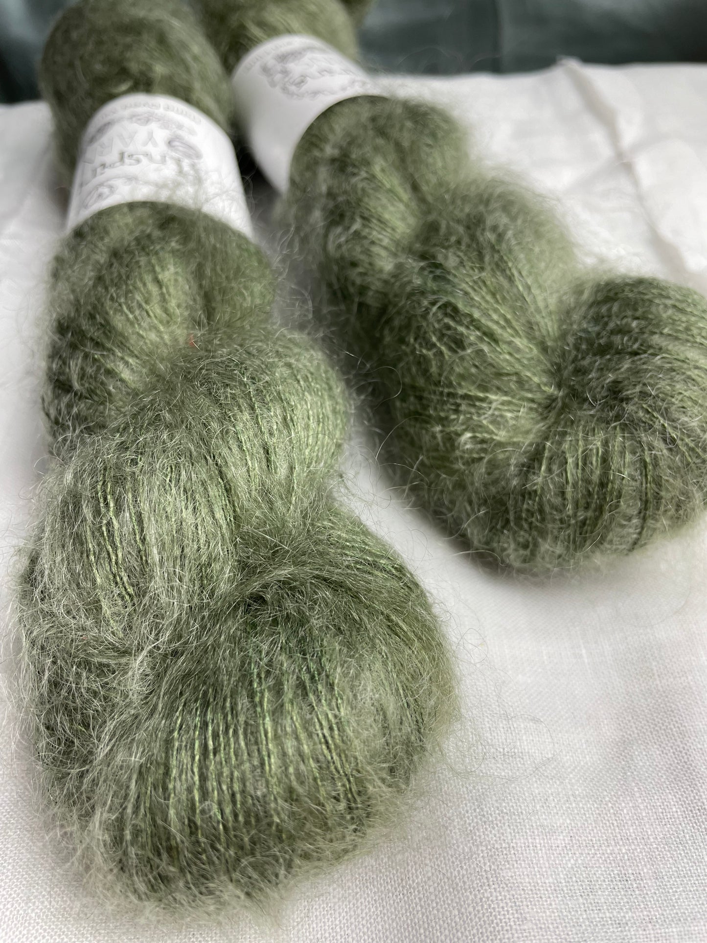 Close up of 2 skeins of light grey green mohair silk lace yarn labelled Inspiring Yarns on a white linen background.