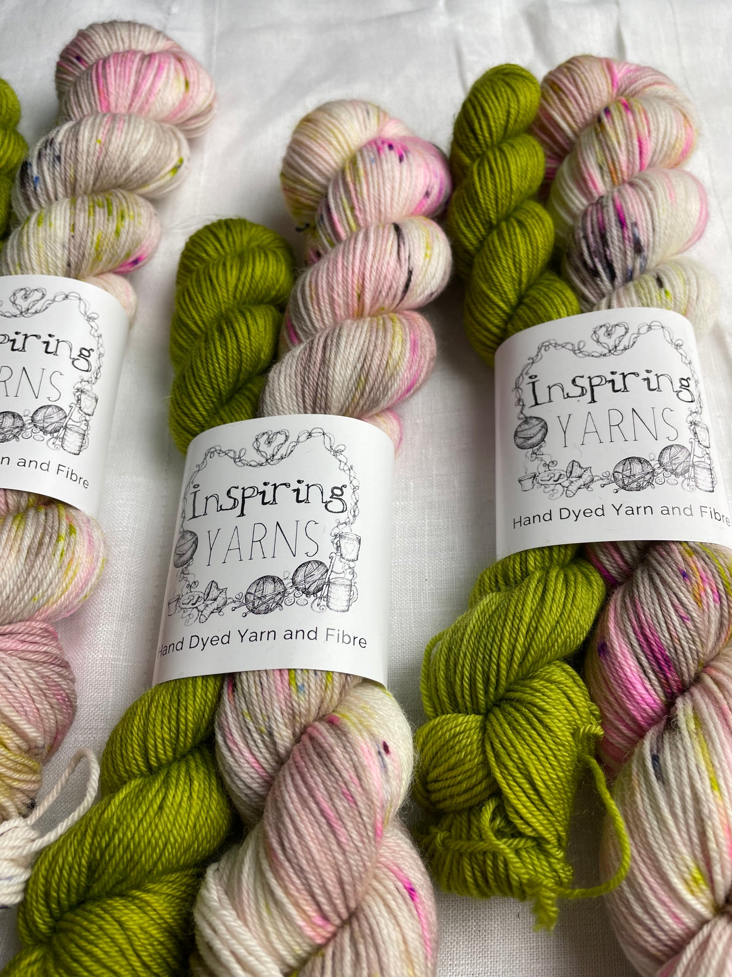Close up of three sets of hand dyed sock yarn comprising one 50g natural speckled and one 20g bright green on a white linen background. The yarn is branded Inspiring Yarns.