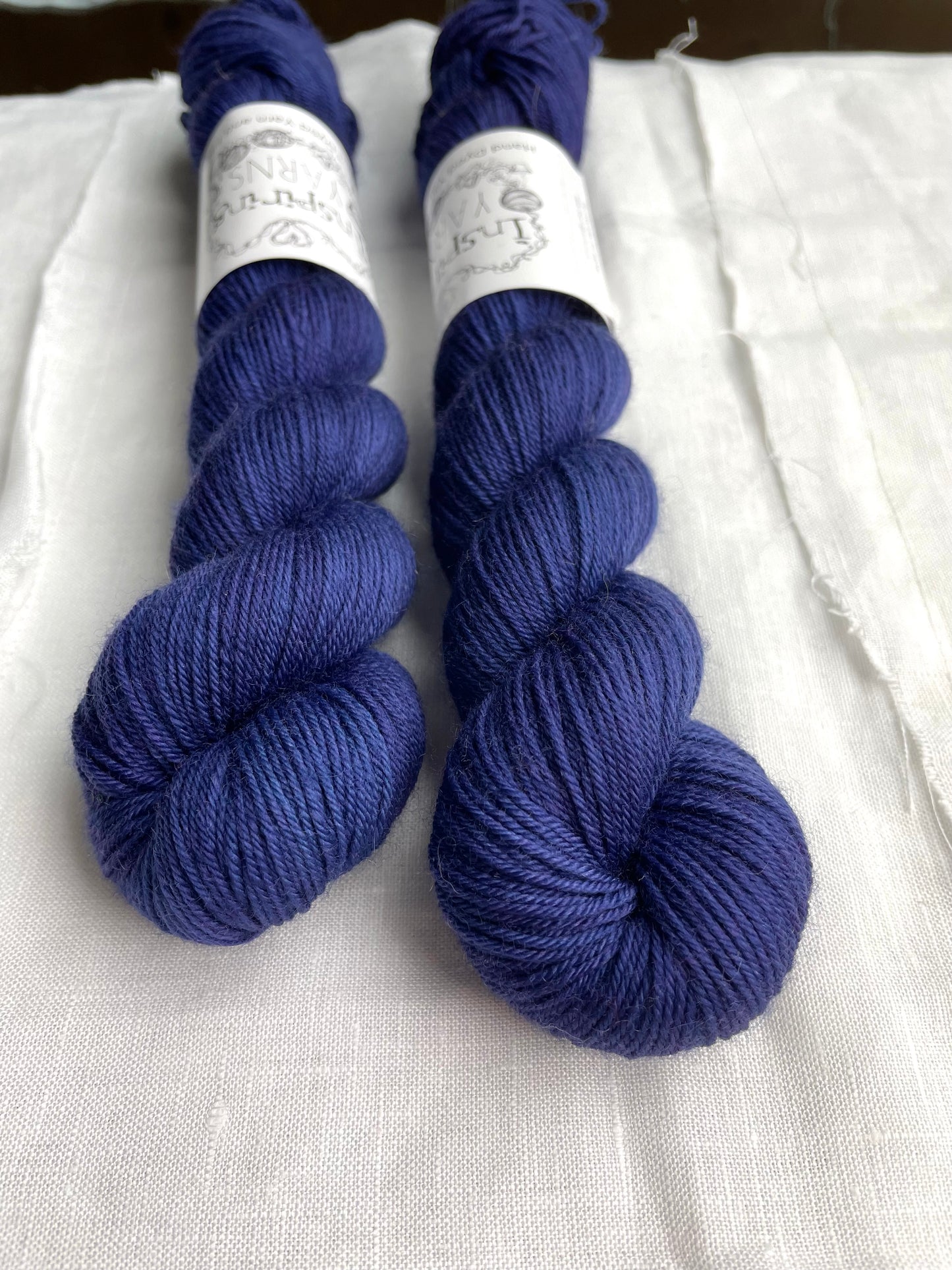 Close up of two tonal deep blue purple coloured skeins of hand dyed Merino Smooth Sock yarn branded Inspiring Yarns on white linen fabric.
