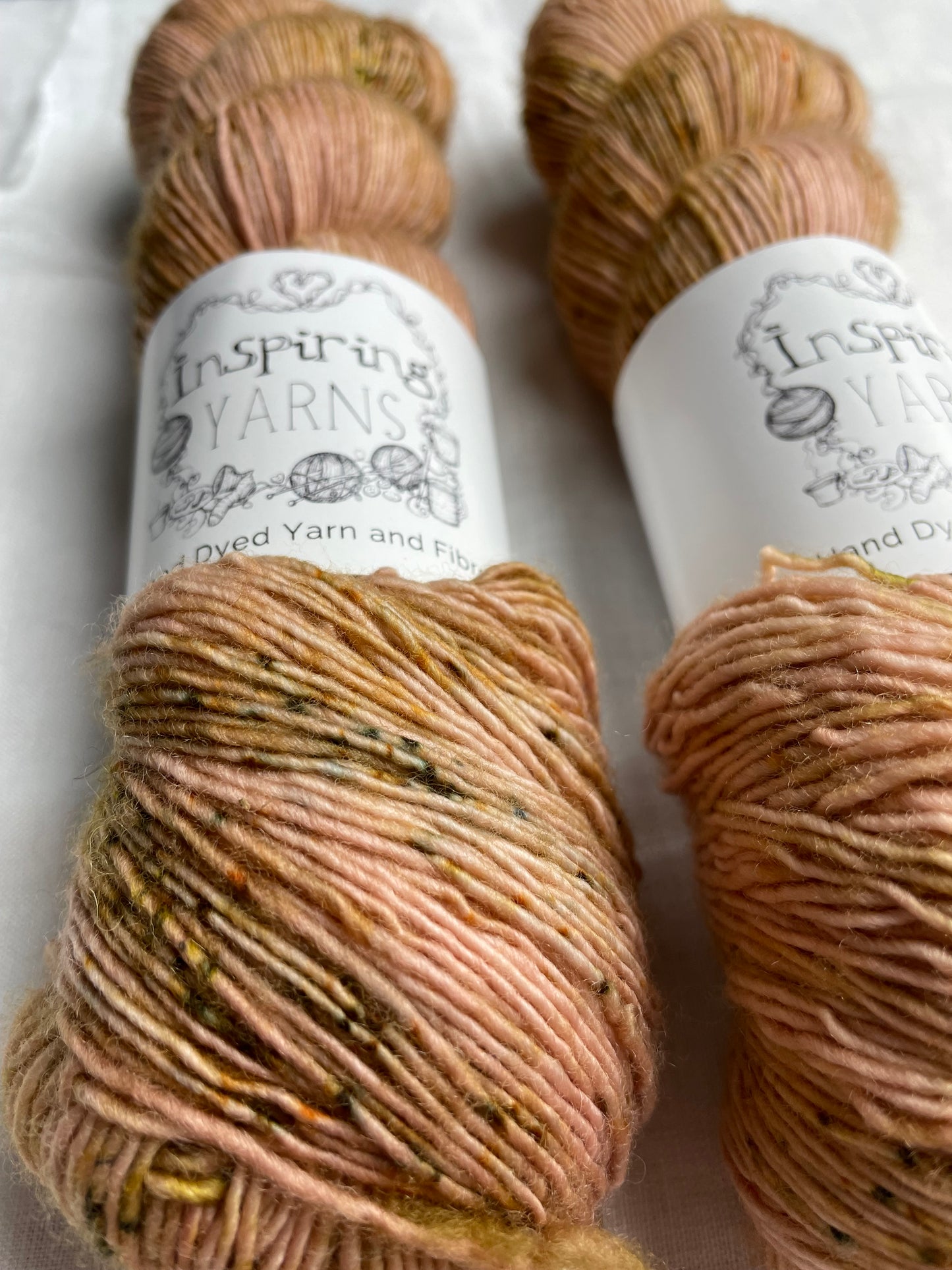 Close up of two skeins of speckled soft terracotta coloured hand dyed Merino Singles yarn labelled Inspiring Yarns on a white linen background.