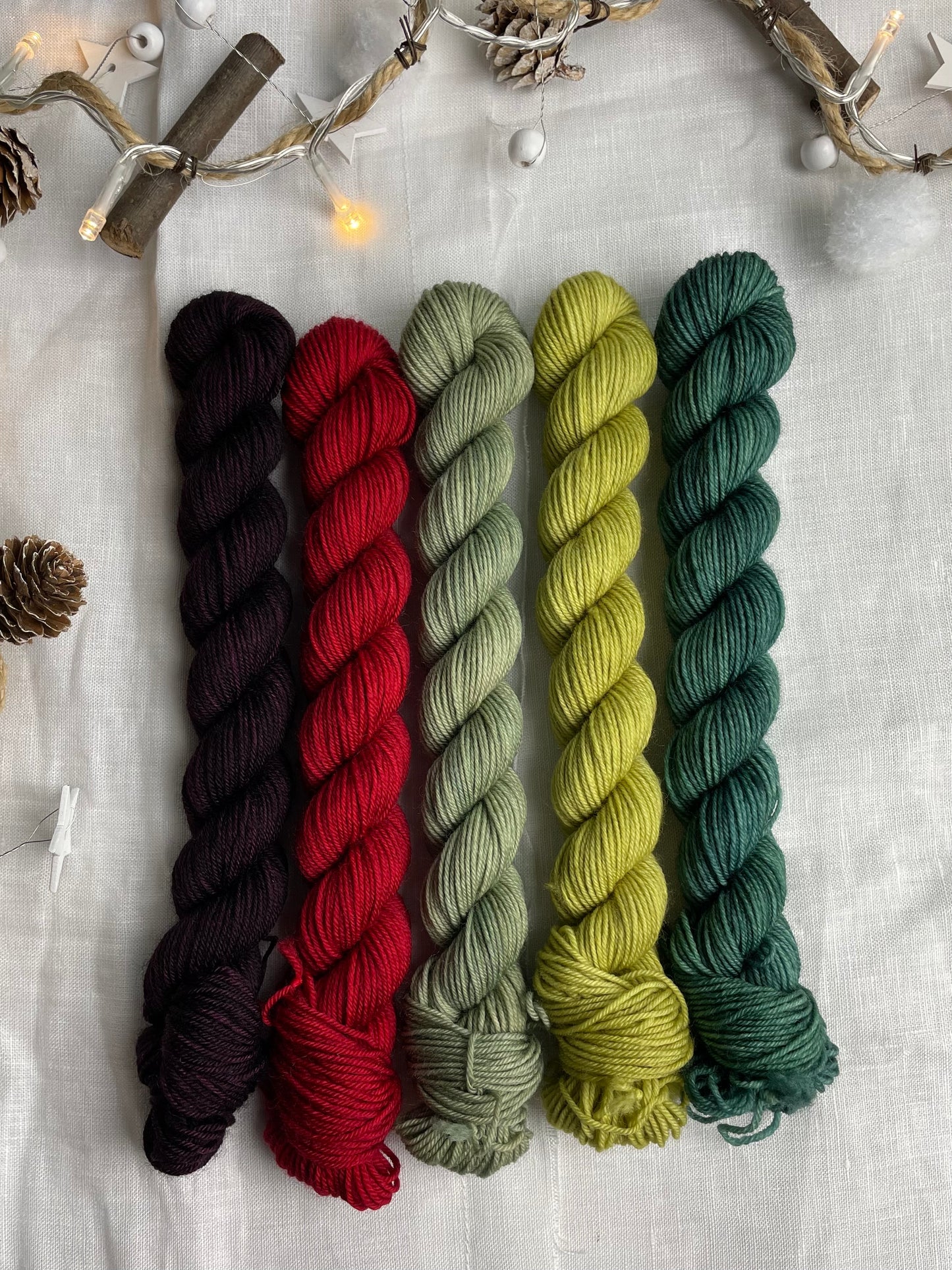 A set of five hand dyed yarn mini skeins on Merino Smooth Sock Yarn in red and green tones labelled Inspiring Yarns. The background is white linen and some Christmas lights with pinecones, twigs and stars.