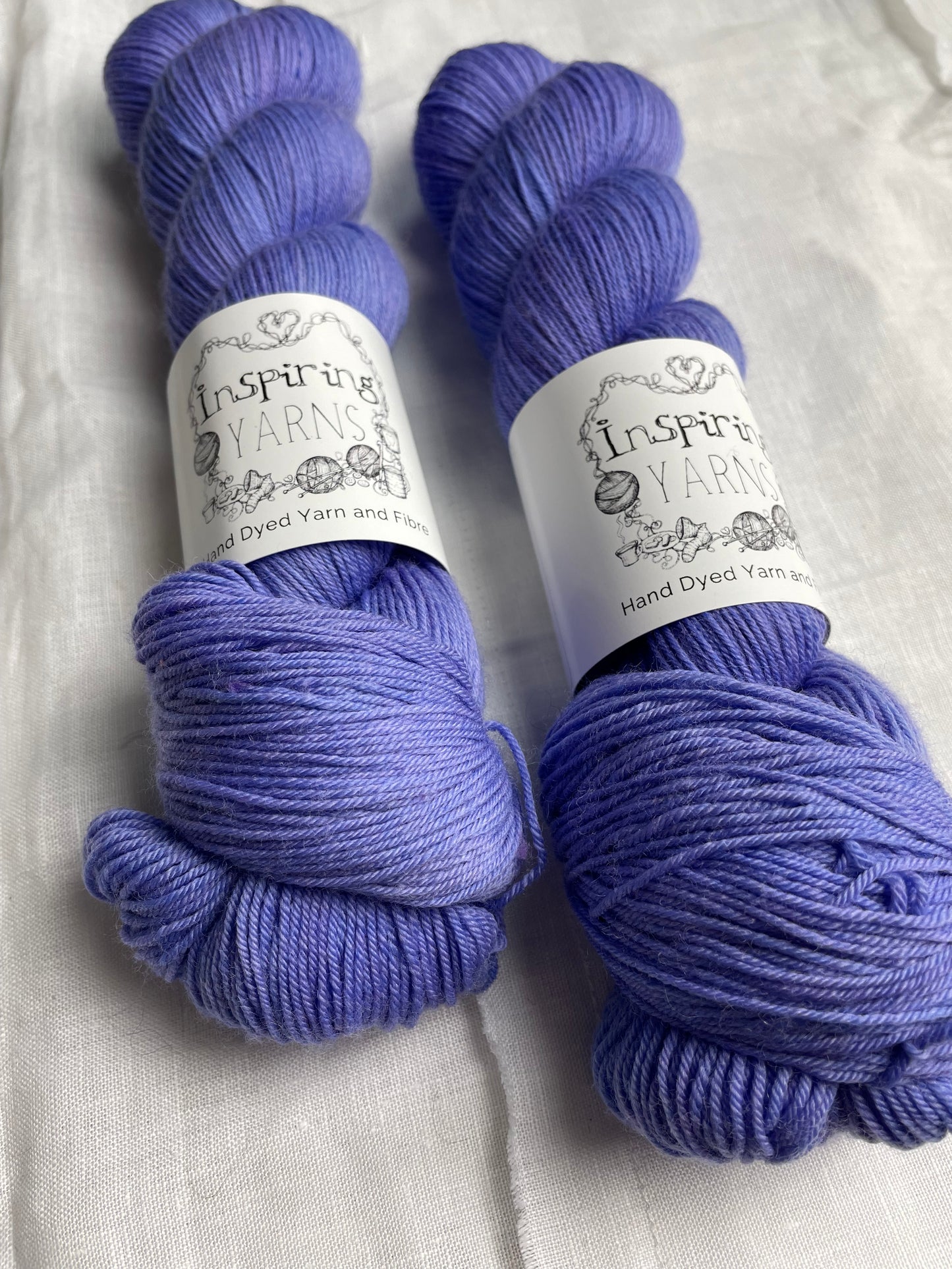 Two skeins of Moonstone, blue lilac coloured Merino Smooth Sock labelled Inspiring Yarns on a white linen background.