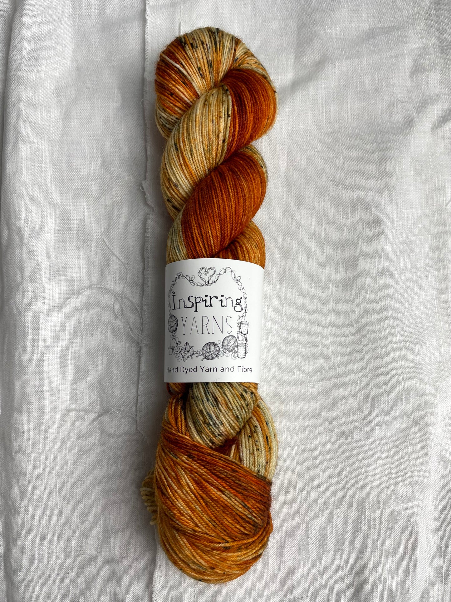 A skein of burnt orange speckled Merino Smooth Sock labelled InspiringYarns lies on a white linen background.