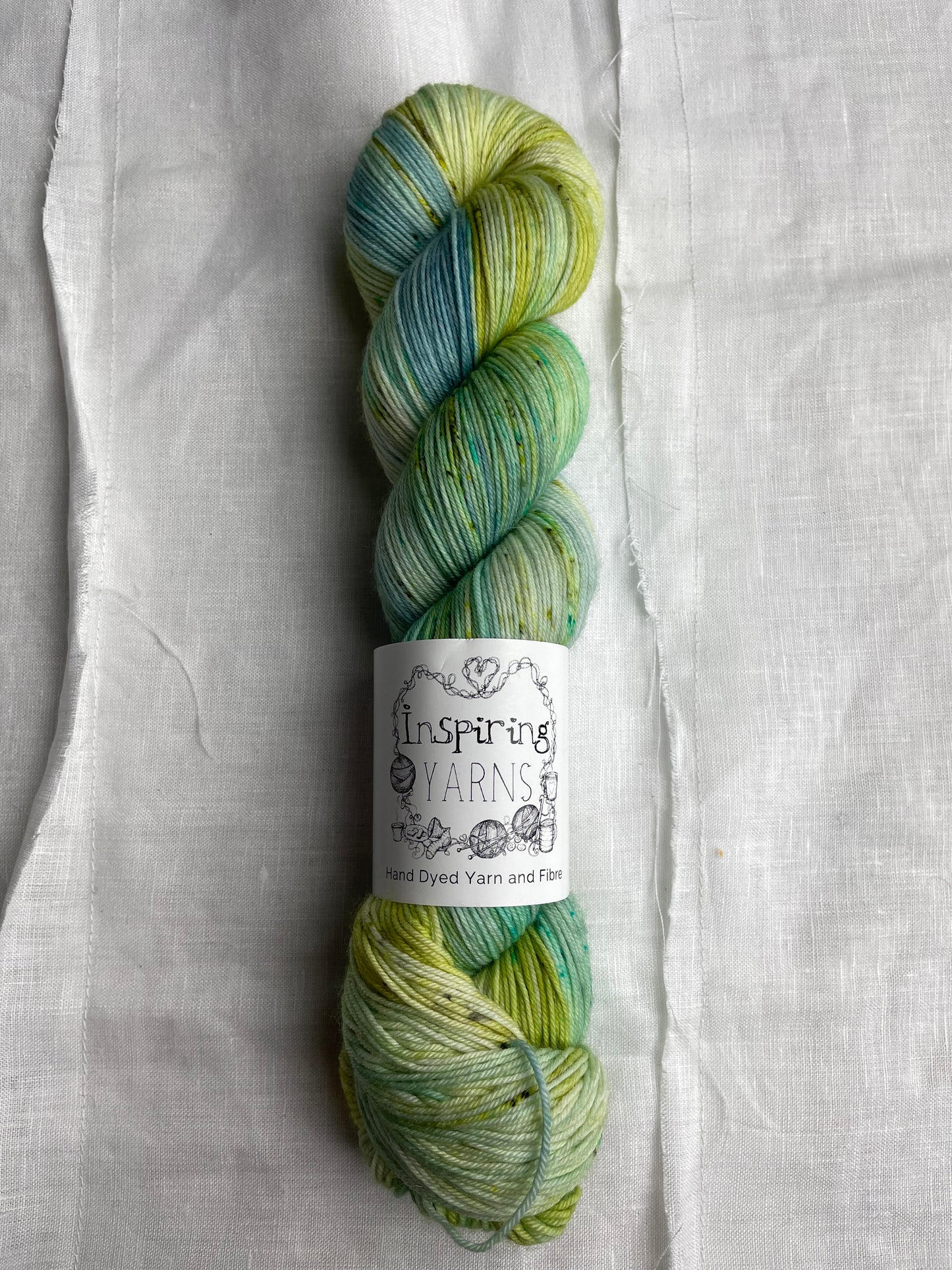 A skein of variegated blue, teal and acid yellow coloured Merino Smooth Sock yarn branded Inspiring Yarns on a white linen fabric background.