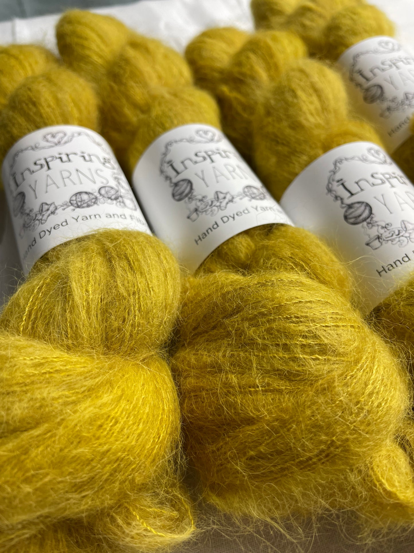 Four skeins of tonal golden ochre coloured hand dyed fluffy Alpaca Silk Lace yarn labelled Inspiring Yarns on a white linen background.