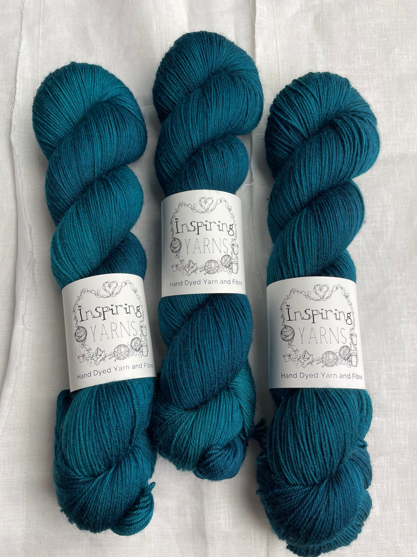 Three intense teal blue coloured skeins of Merino Smooth Sock yarn branded Inspiring Yarns on a white linen fabric background.