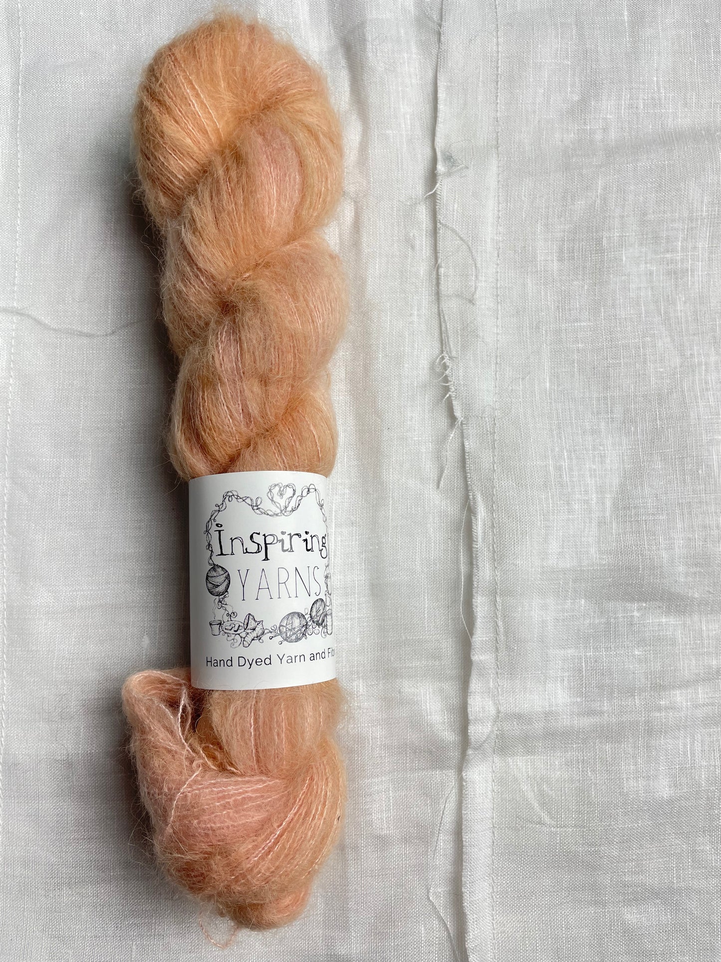 A skein of dusky peach coloured hand dyed fluffy Alpaca Silk Lace yarn labelled Inspiring Yarns on a white linen background.