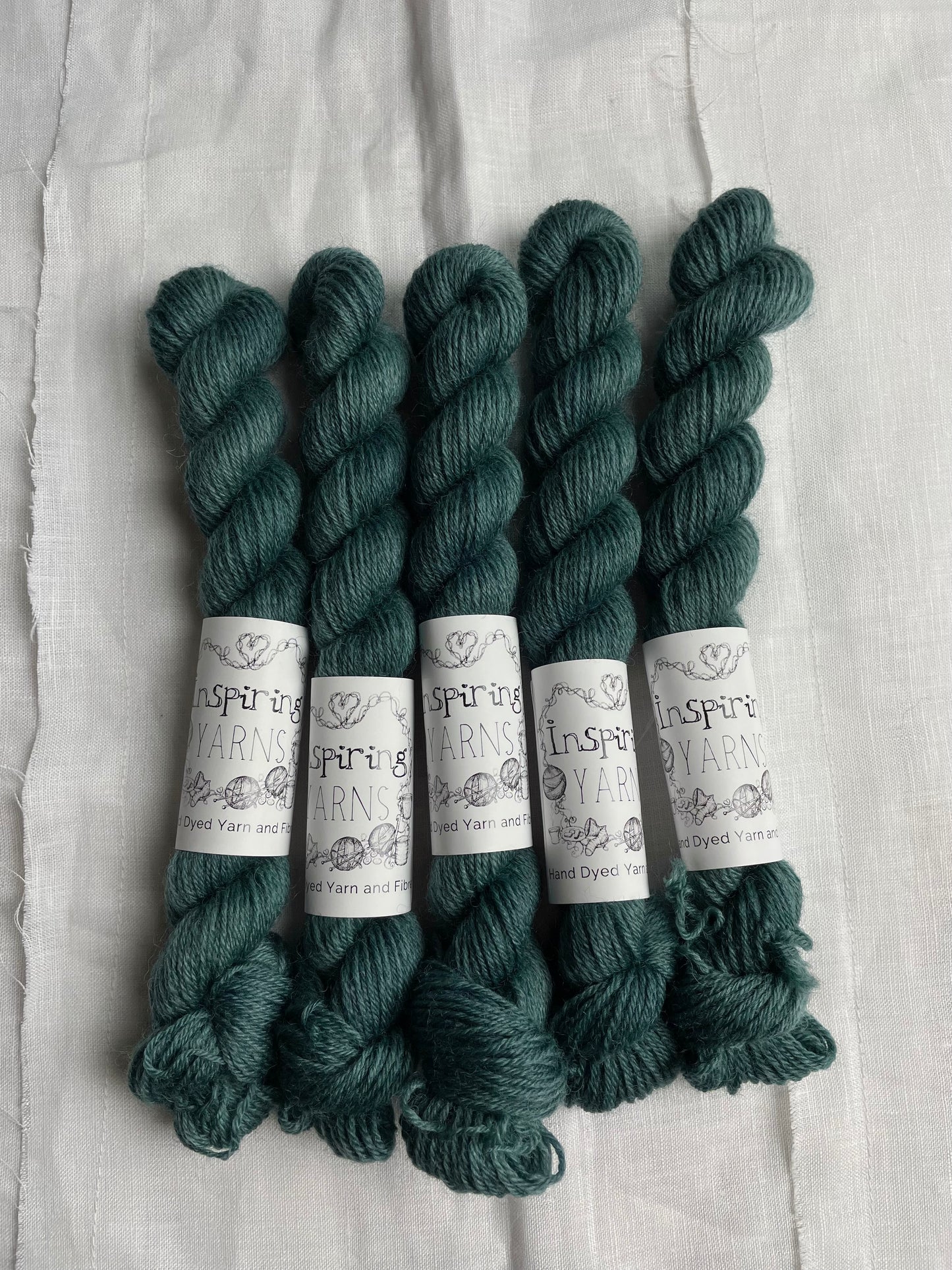 Five mini skeins of a tonal dark teal Highland 4Ply yarn branded Inspiring Yarns on a white linen background.