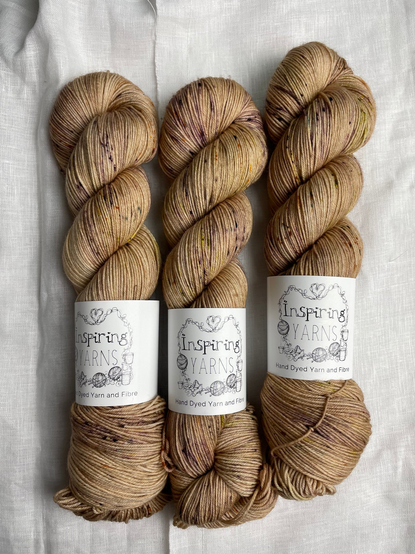 Three skeins of Harvest, a pale gold with speckled coloured Merino Smooth Sock, labelled Inspiring Yarns on a white linen background.