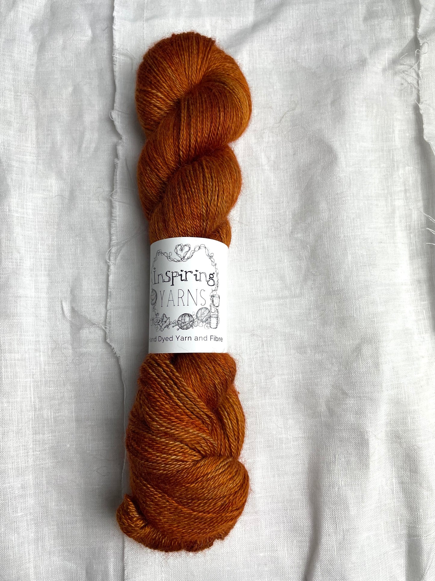 A skein of burnt orange mist 4ply yarn labelled Inspiring Yarns on a white linen background.