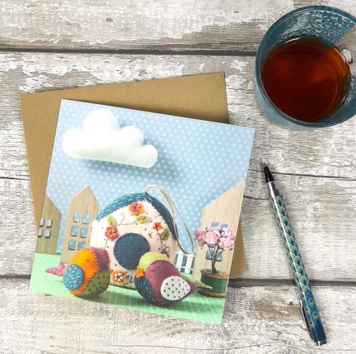 Greeting card with colourful house illustration on an aged wooden surface with a cup of tea and pen.