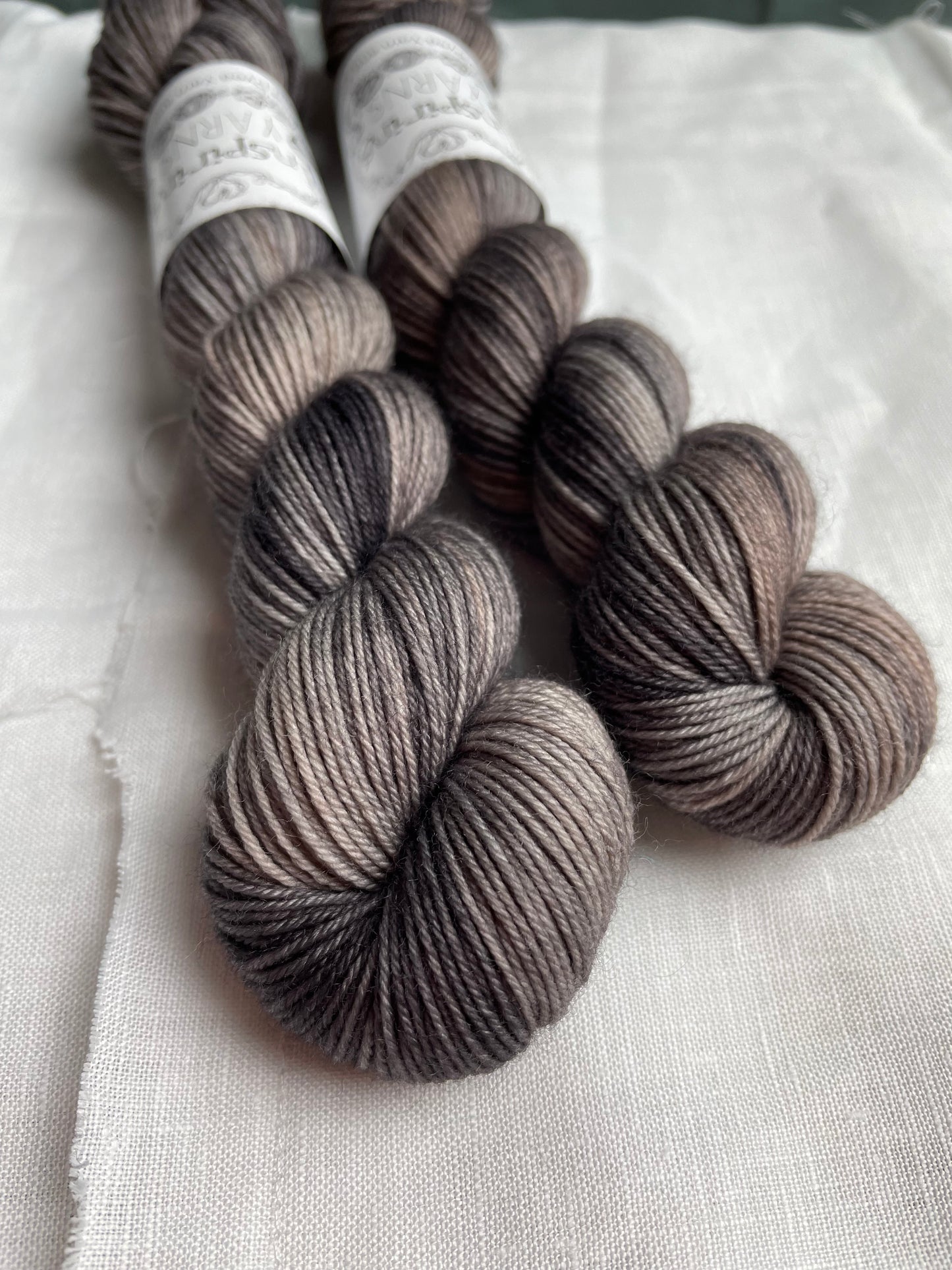 Two skeins of Merino Smooth Sock yarn hand dyed in a tonal grey colourway and labelled Inspiring Yarns on a white linen background.