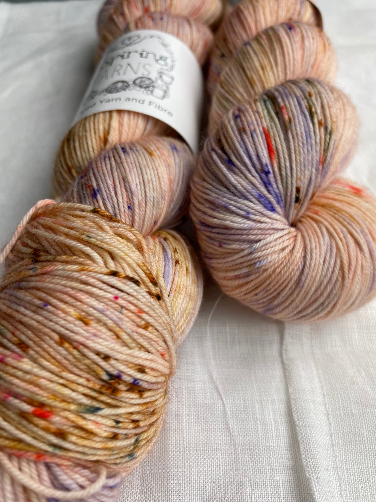 Close up of two speckled peach coloured skeins of Merino Smooth Sock yarn branded Inspiring Yarns on a white linen fabric background.