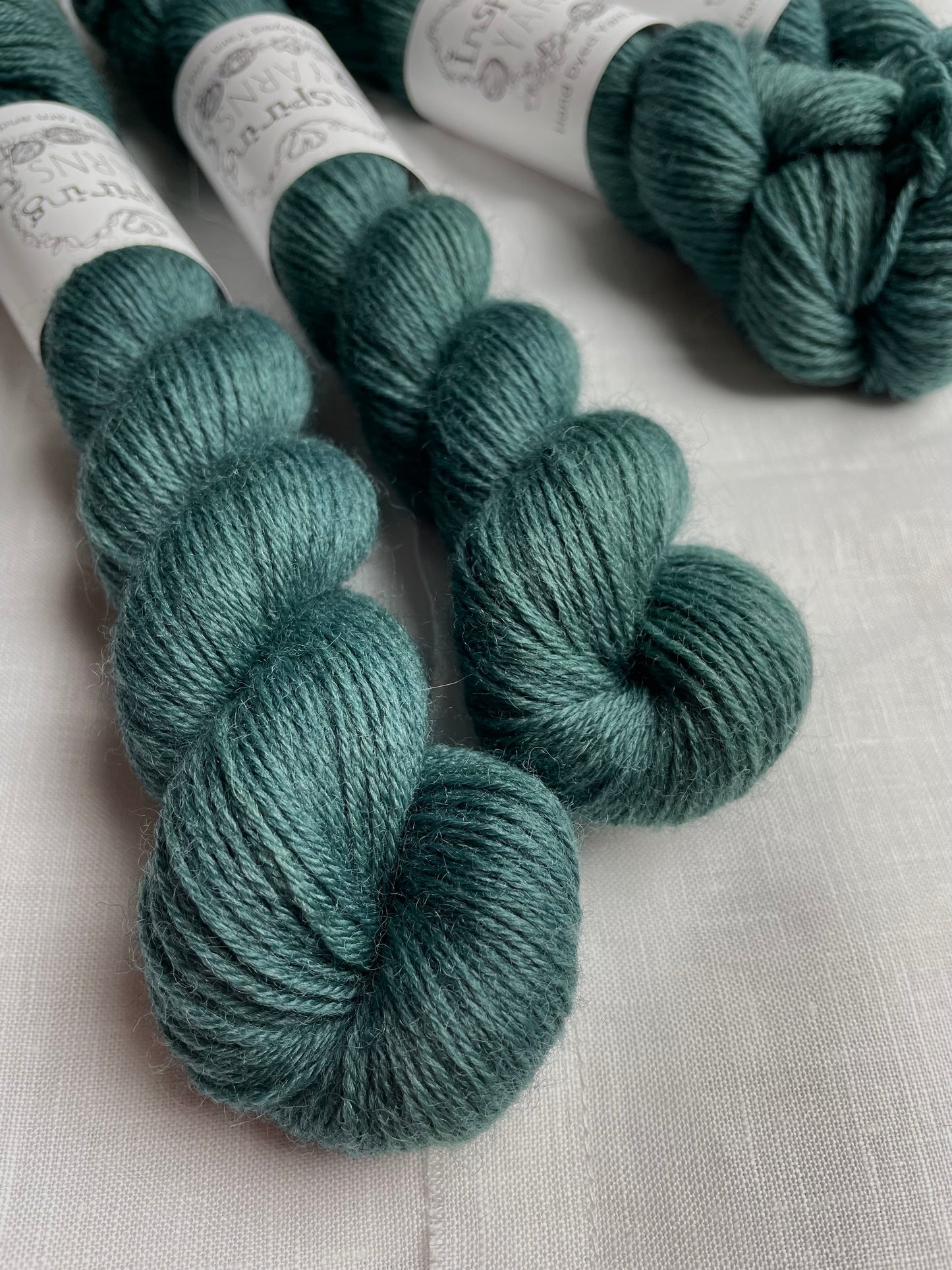 Four skeins of a tonal dark teal Highland 4Ply yarn branded Inspiring Yarns on a white linen background.