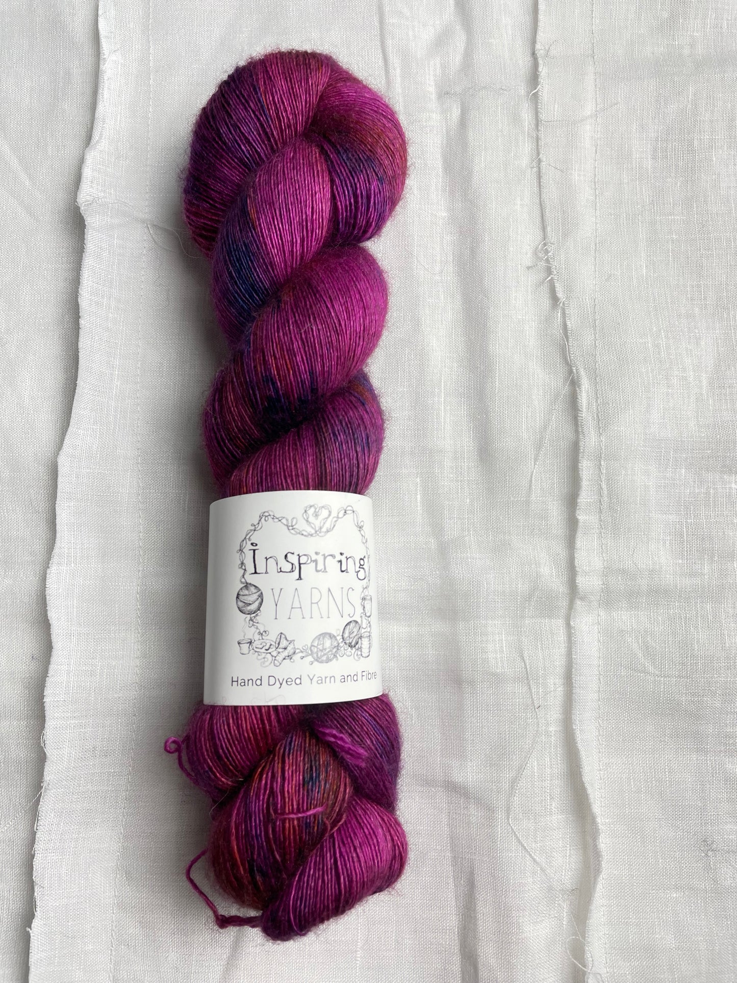 A speckled fuchsia pink coloured skeins of hand dyed Yak Silk Singles branded Inspiring Yarns on a white linen background.