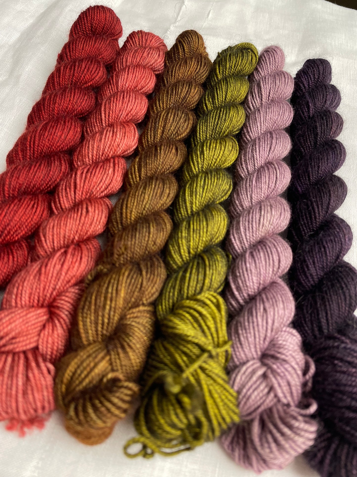 Close up of a set of 6 mini skeins of various colours of Yak Sock yarn on a white linen background.