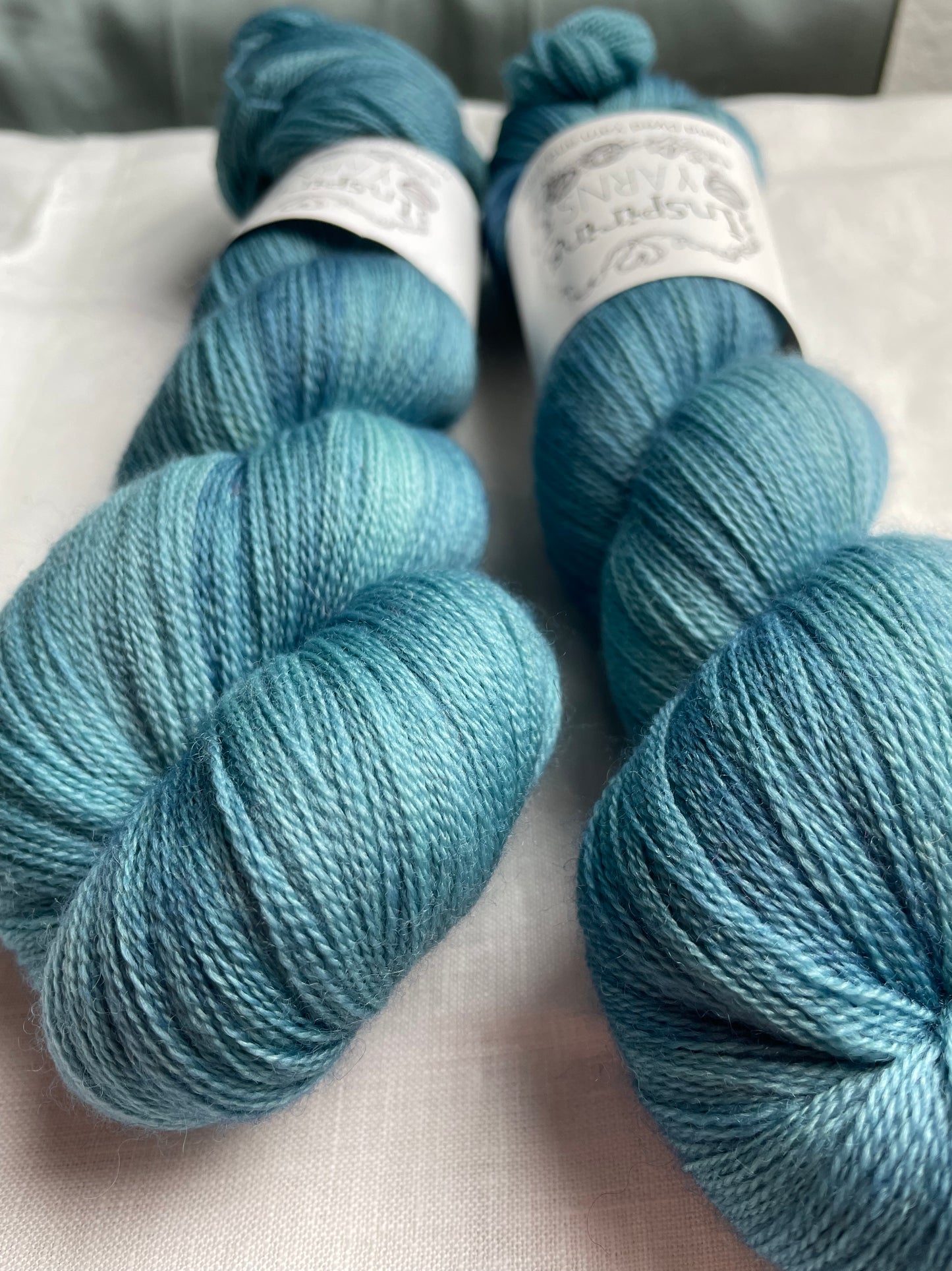 Close up of two skeins of tonal soft teal coloured hand dyed Merino Lace yarn branded Inspiring Yarns on a white linen cloth.