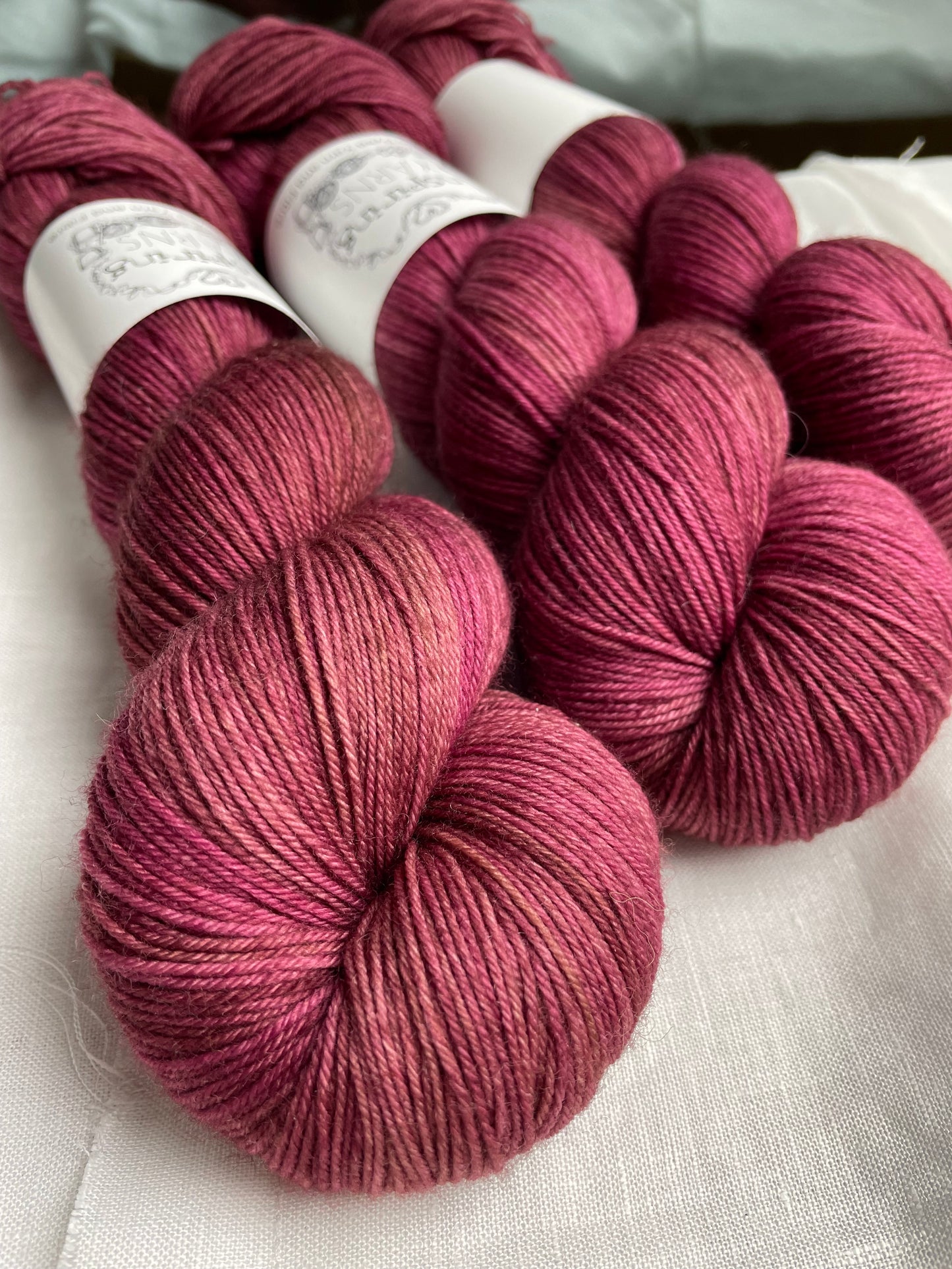 Close up of three skeins of dusky pink Merino Smooth Sock yarn labelled Inspiring Yarns on a white linen background.