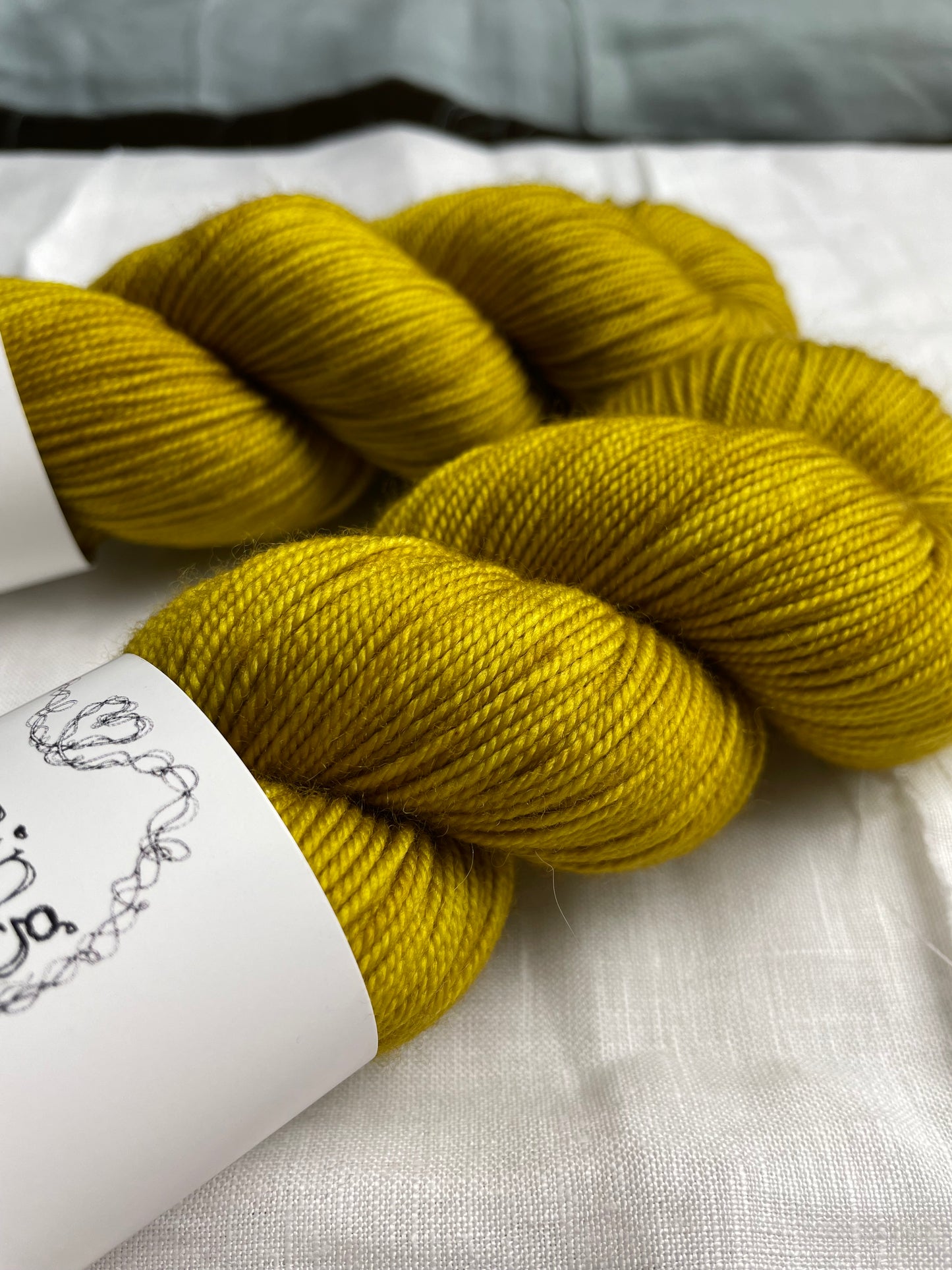 Close up of two skeins of tonal golden ochre coloured hand dyed Merino Sport yarn branded Inspiring Yarns on a background of white linen.