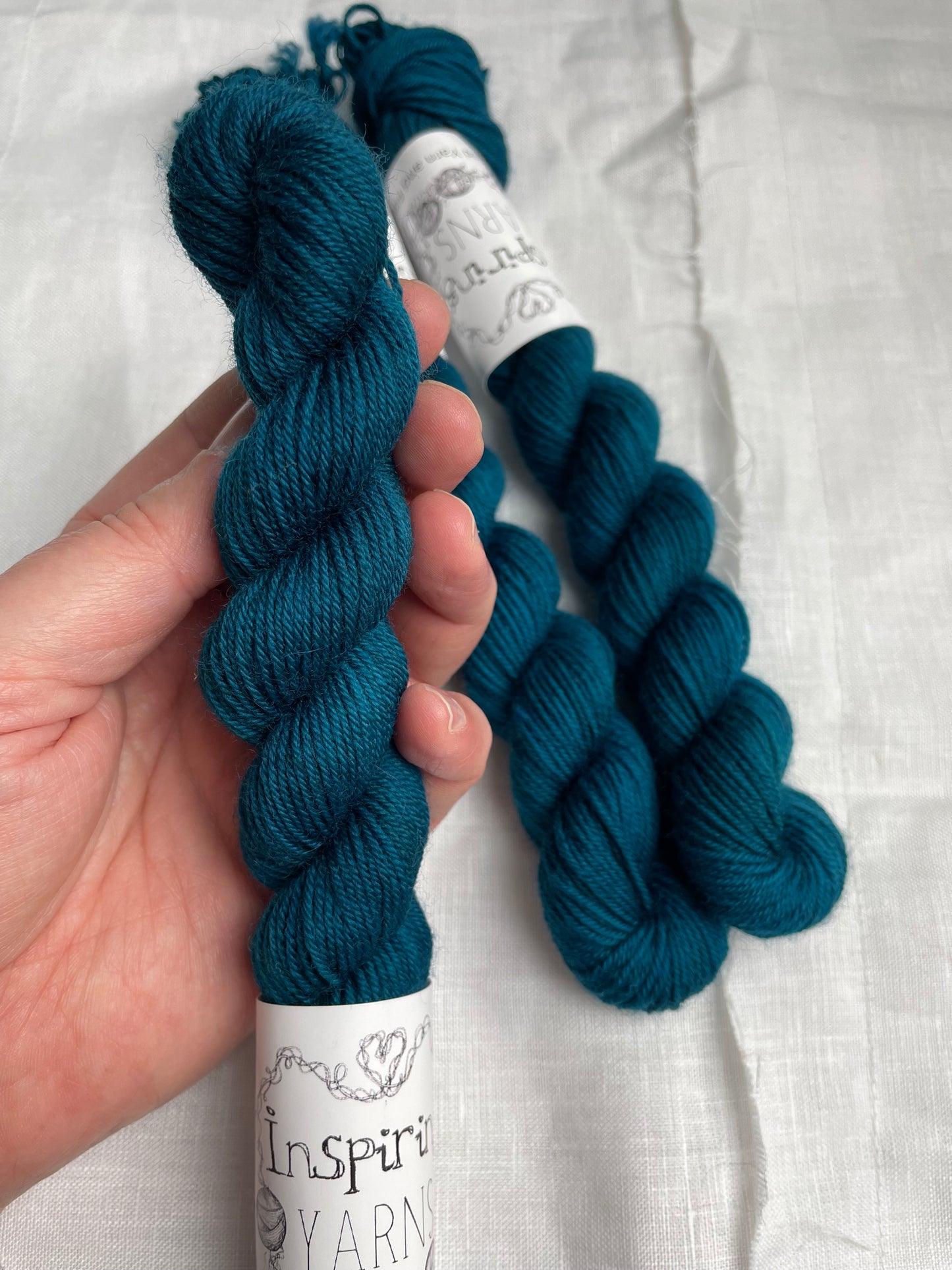 Hand holding an intense teal blue coloured skein of Merino Smooth Sock yarn branded Inspiring Yarns in front of a white linen fabric background. Two more skeins lie on the linen.