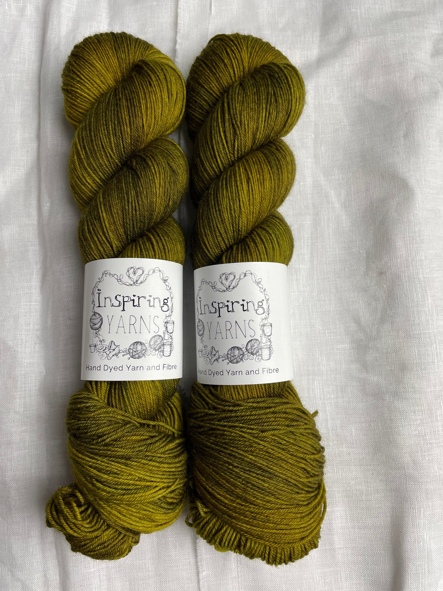 Two skeins of Gremlin, a dark olive green coloured Merino Smooth Sock, labelled Inspiring Yarns on a white linen background.