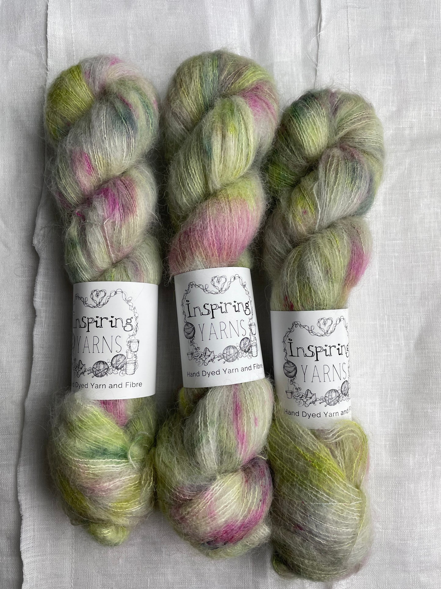 Three skeins of green and pink speckled silver grey coloured hand dyed fluffy Alpaca Silk Lace yarn labelled Inspiring Yarns on a white linen background.