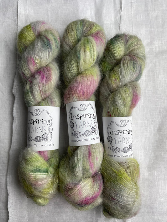 Three skeins of green and pink speckled silver grey coloured hand dyed fluffy Alpaca Silk Lace yarn labelled Inspiring Yarns on a white linen background.