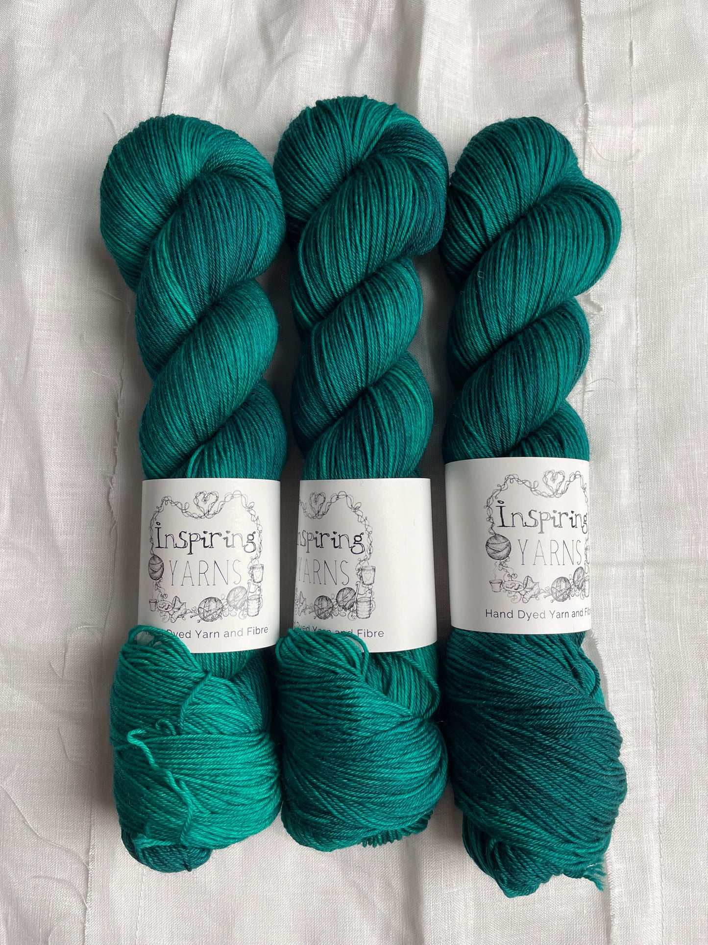 Three skeins of Merino Smooth Sock yarn hand dyed in a tonal jade green colourway and labelled Inspiring Yarns on a white linen background.