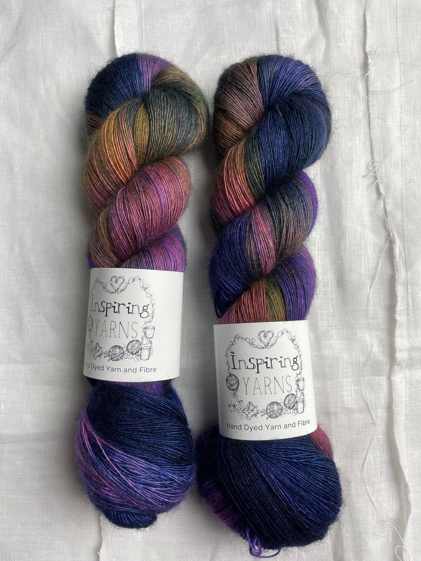Two variegated multicoloured skeins of hand dyed Yak Silk Singles branded Inspiring Yarns on a white linen background.