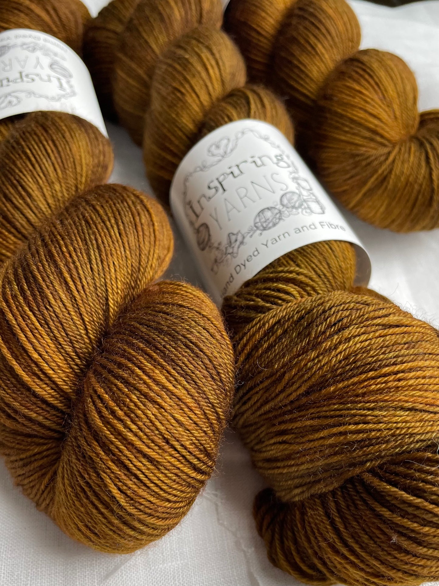 Three skeins of Kelp, a bronze tonal coloured Merino Smooth Sock, labelled Inspiring Yarns on a white linen background.