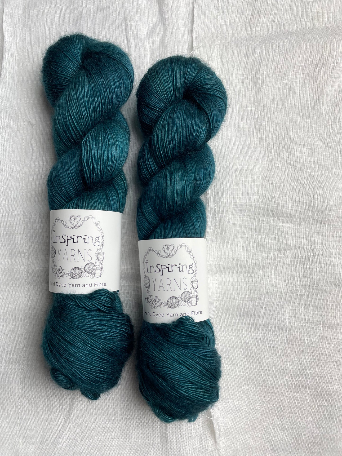 Two tonal intense teal blue coloured skeins of hand dyed Yak Silk Singles branded Inspiring Yarns on a white linen background.