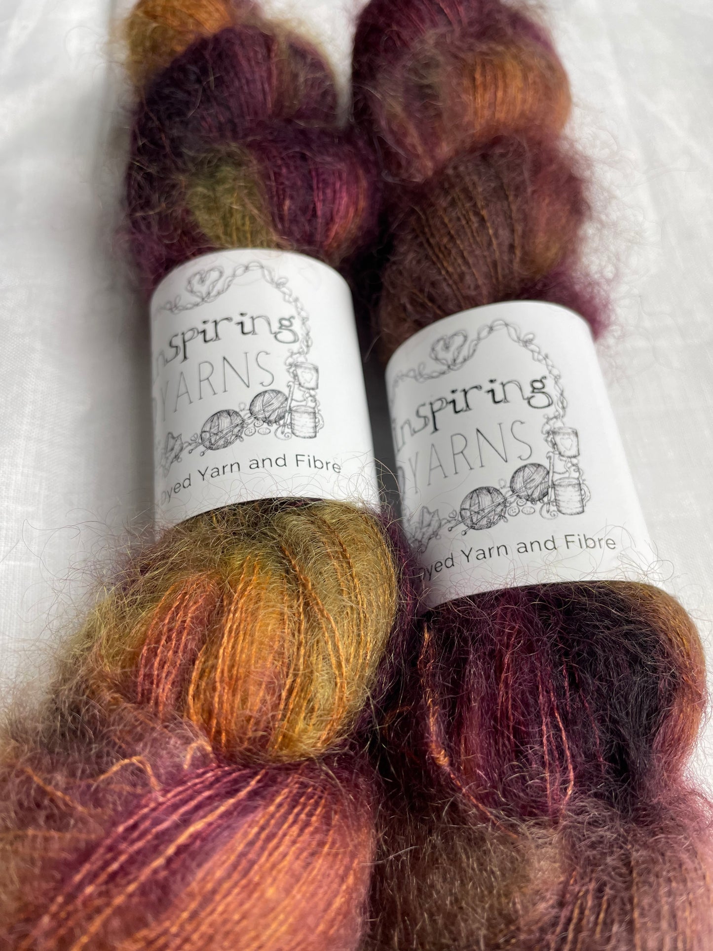 Close up of 2 skeins of variegated tan burgundy and green mohair silk lace yarn labelled Inspiring Yarns on a white linen background.