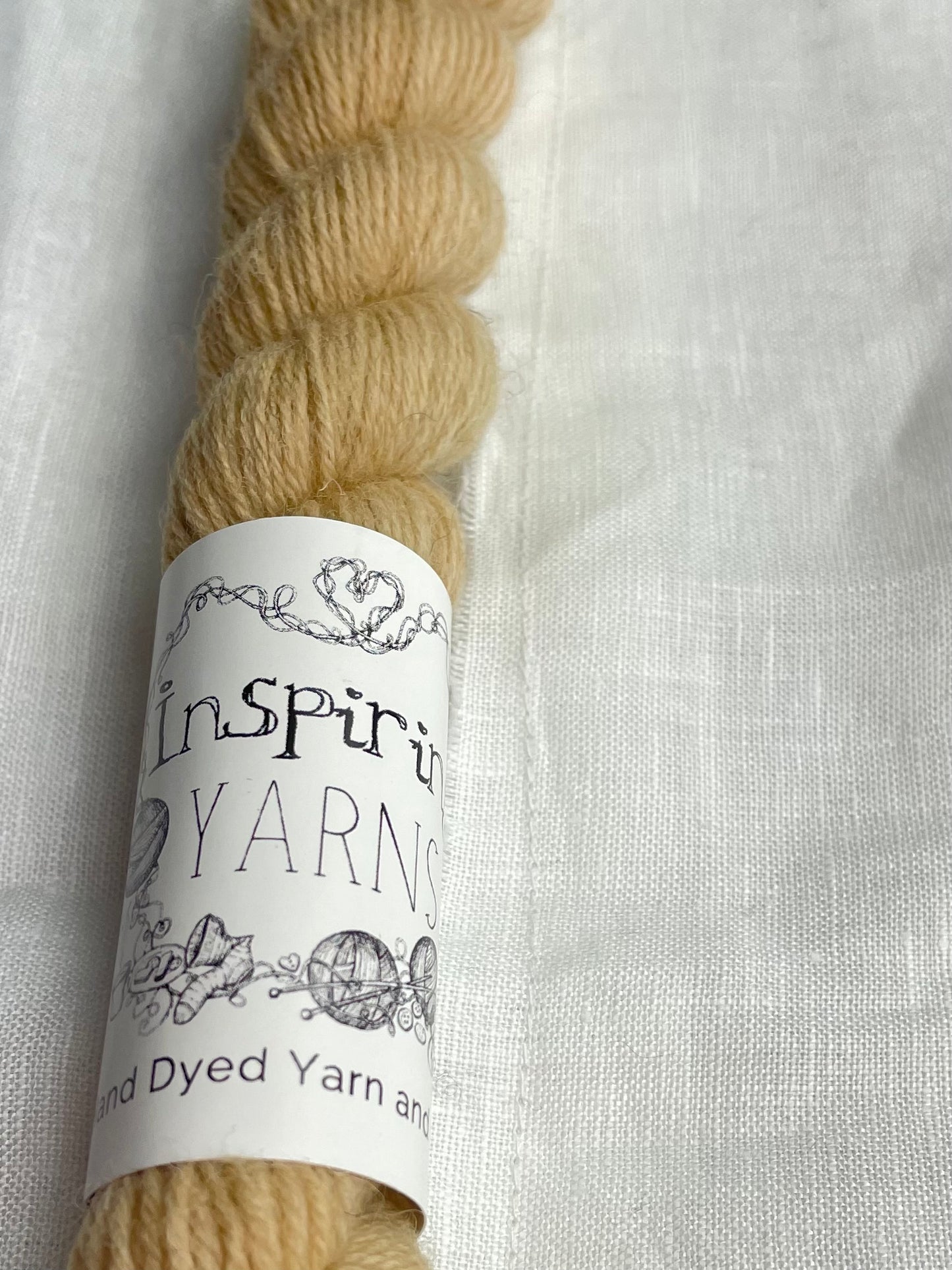 A close up of a mini skein of tonal golden caramel coloured Highland 4Ply yarn branded Inspiring Yarns on a white linen background.
