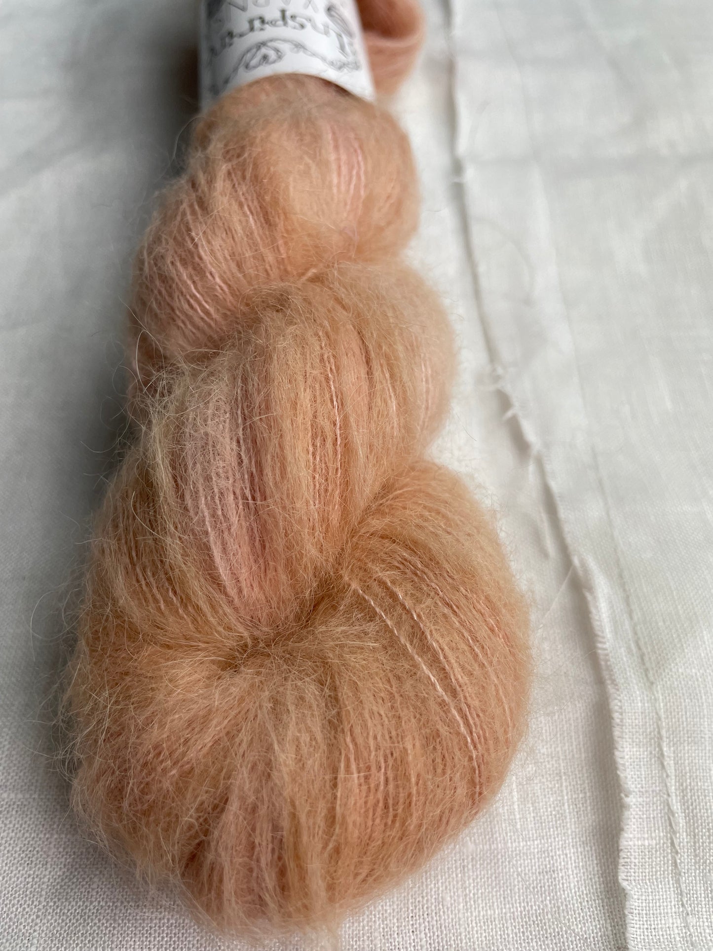 Close up of a skein of dusky peach coloured hand dyed fluffy Alpaca Silk Lace yarn labelled Inspiring Yarns on a white linen background.