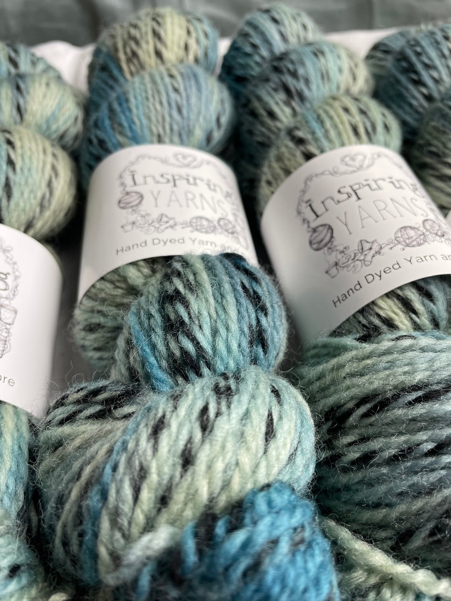 Close up of handyed Highland Zebra yarn in light green and teal blue colourway. The yarn has black strands spun in to it. The skeins are labelled as Inspiring Yarns.