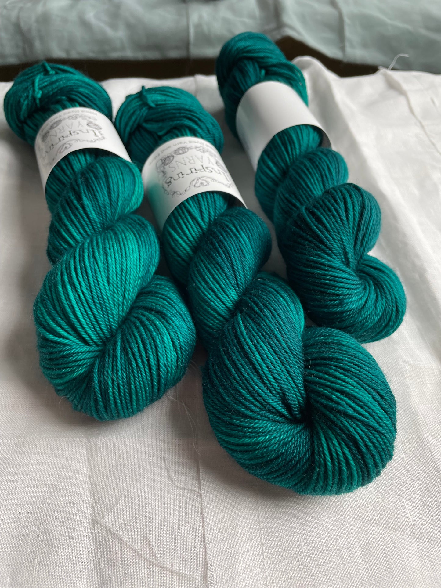Three skeins of Merino Smooth DK yarn hand dyed in a tonal jade green colourway and labelled Inspiring Yarns on a white linen background.