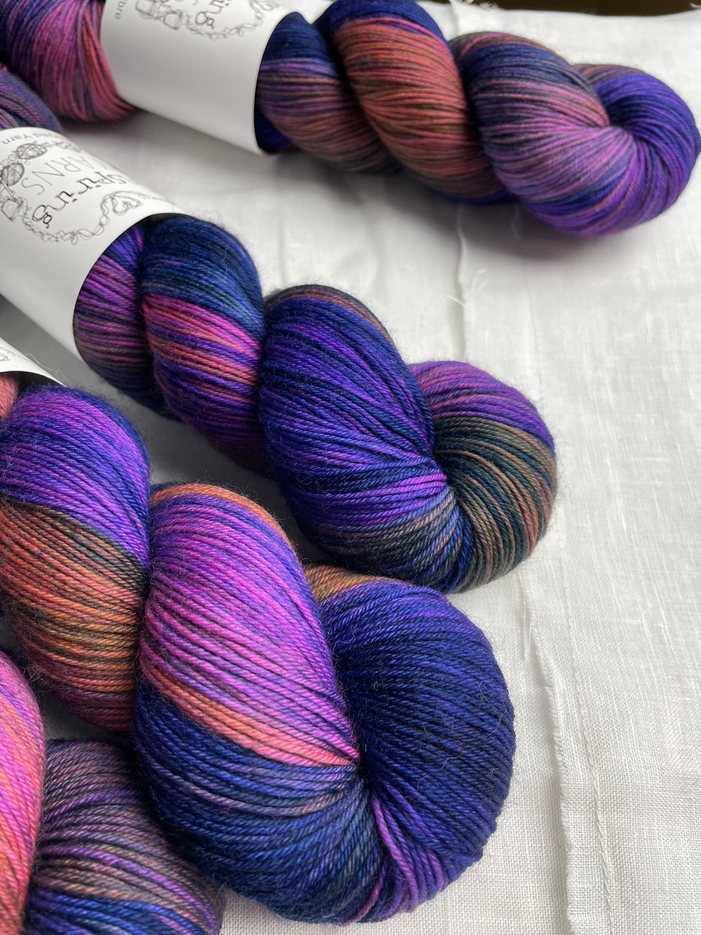 Close up of four variegated blue and multi coloured skeins of Merino Smooth Sock yarn branded Inspiring Yarns on a white linen fabric background.