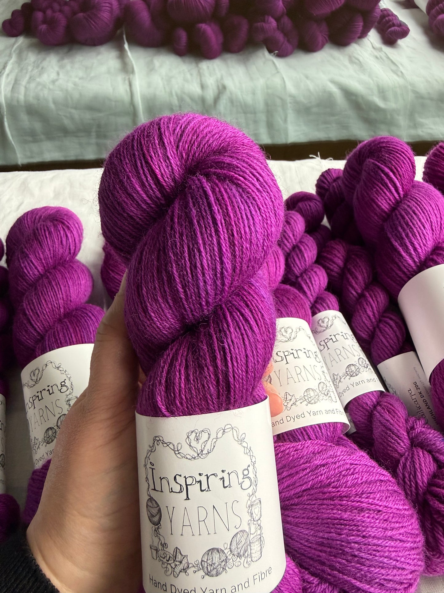 Hand holding a skein of fuchsia tonal yarn labelled Inspiring Yarns. More skeins of yarn of the same colour sit in the background.