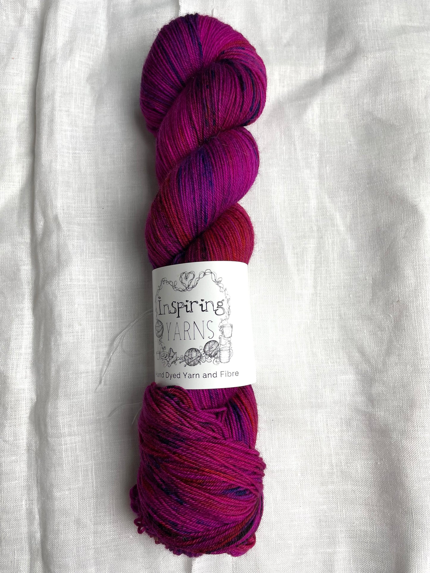 A skein of fuchsia pink speckled Merino Smooth Sock labelled InspiringYarns lies on a white linen background.