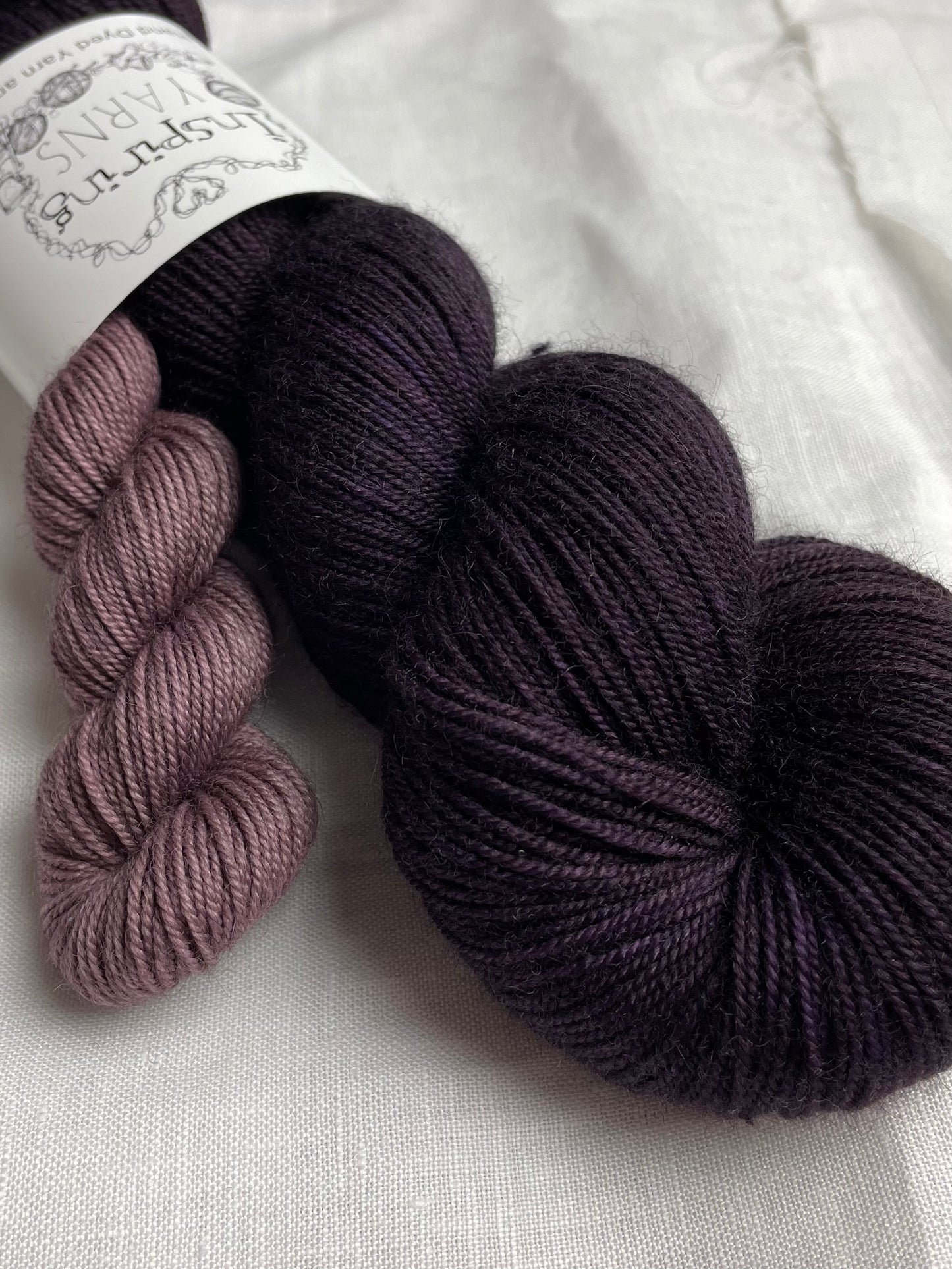 Close up of a sock set made of a skein of dark tonal purple hand dyed yarn with a contrasting dusty lilac mini skein, labelled Inspiring Yarns and laid on a white linen cloth.